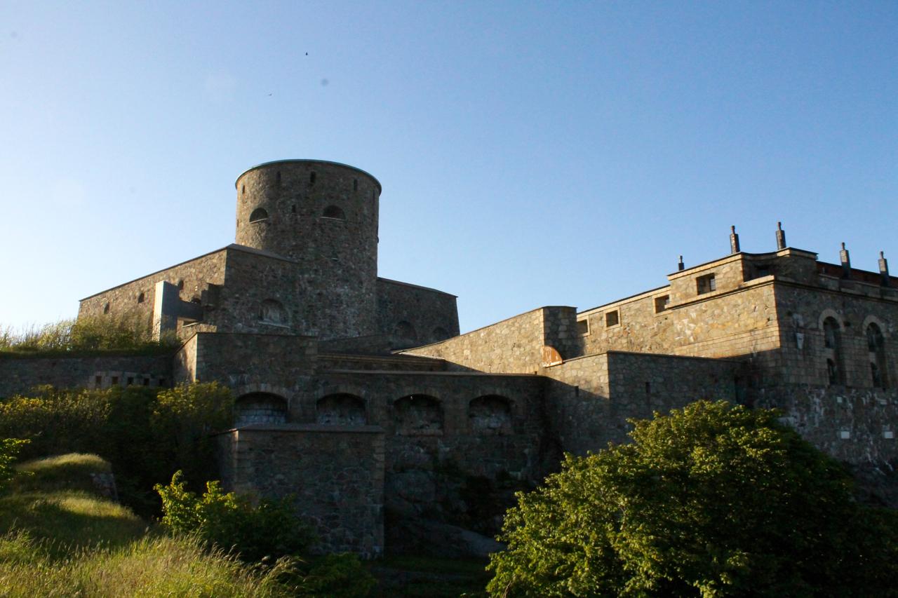A stone fortress with a round tower and multiple levels, surrounded by greenery and under a clear blue sky. Marstrand Island Trail System mountain bike trail.