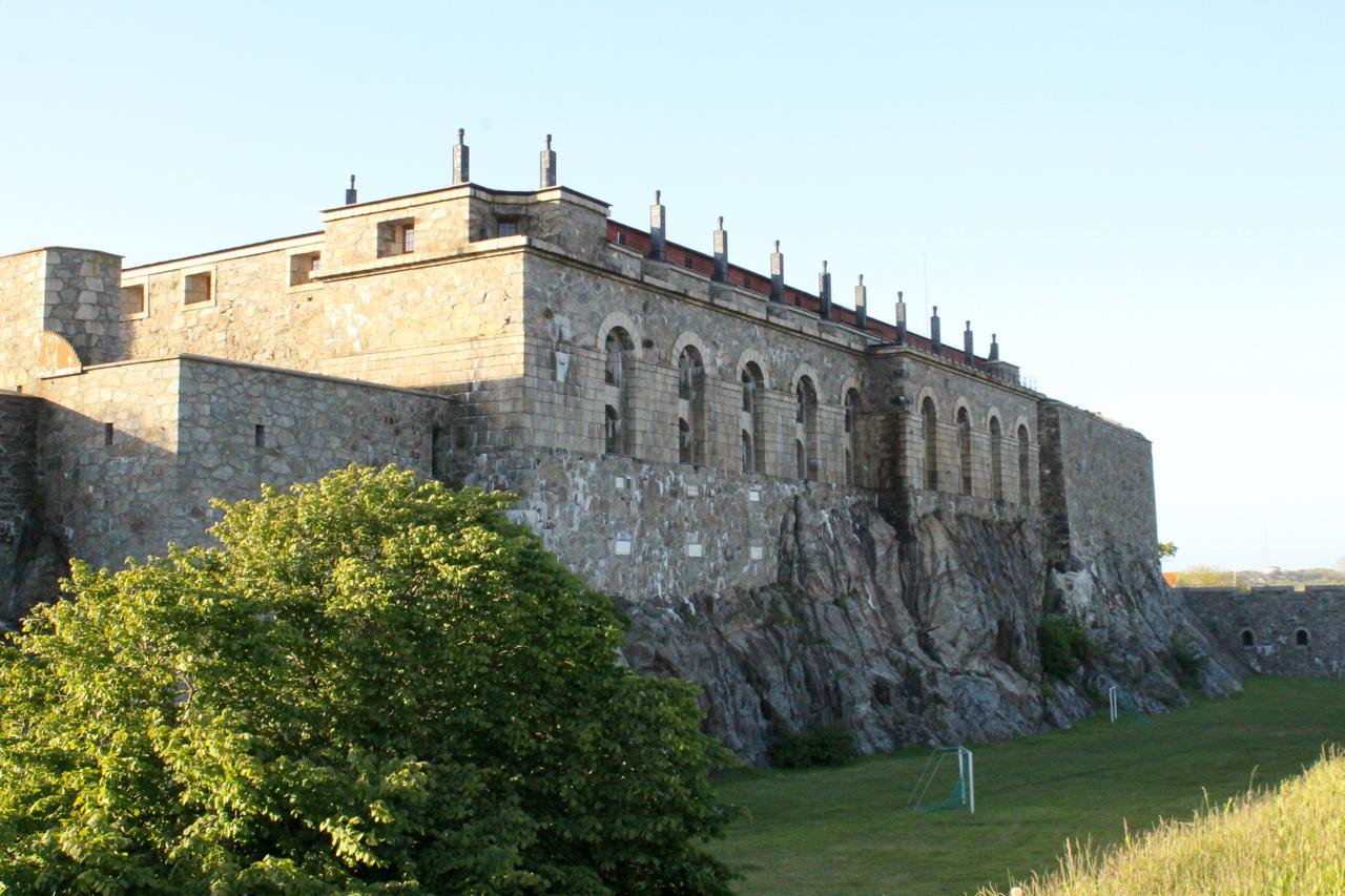 A historical stone fortress built into a rocky hillside, featuring arched windows and battlements along the roof. Lush green trees and grass are visible in the foreground, with clear blue skies in the background. Marstrand Island Trail System mountain bike trail.