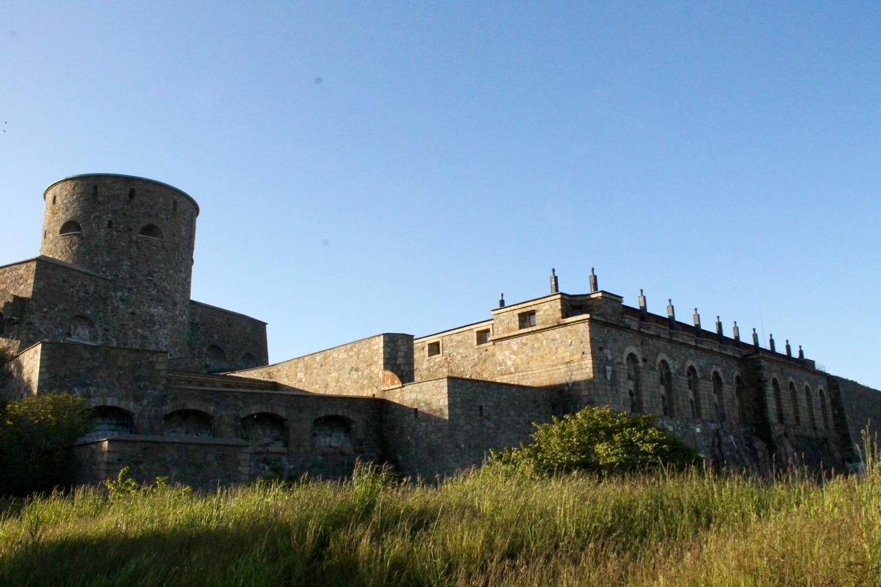 A historic stone fortress with a cylindrical tower, set against a clear blue sky. The structure features multiple arched windows and is partially surrounded by grassy terrain. Marstrand Island Trail System mountain bike trail.