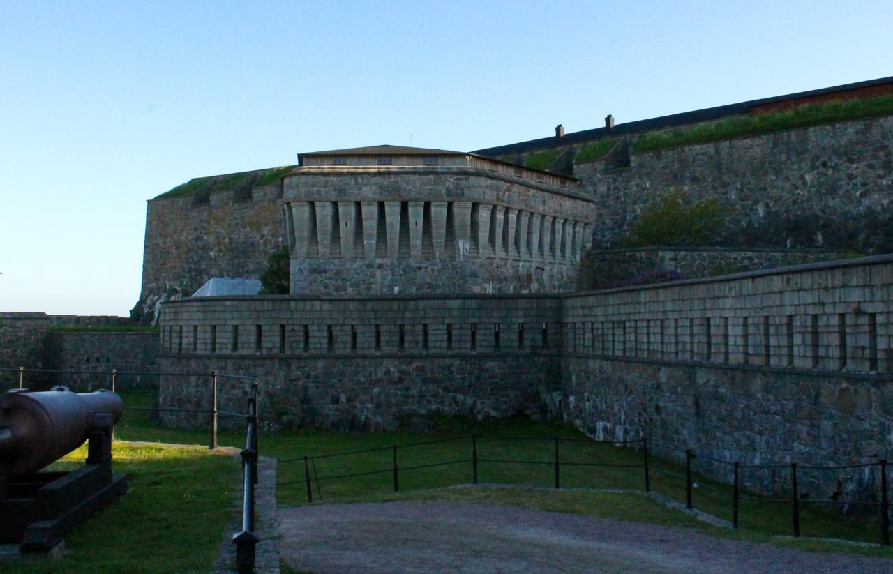A historical stone fortification with towering walls, featuring a round structure with columns on one side. In the foreground, there is an old cannon positioned on the grass, surrounded by a low fence. The sky is clear and bright, indicating early morning or late afternoon light. Marstrand Island Trail System mountain bike trail.