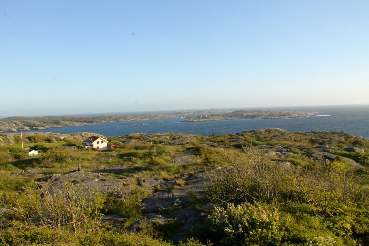 A scenic view of a coastal landscape featuring rocky terrain, lush greenery, and a distant view of the ocean with islands in the background. In the foreground, two small houses are nestled among the hills, providing a picturesque contrast to the expansive blue water under a clear sky. Marstrand Island Trail System mountain bike trail.