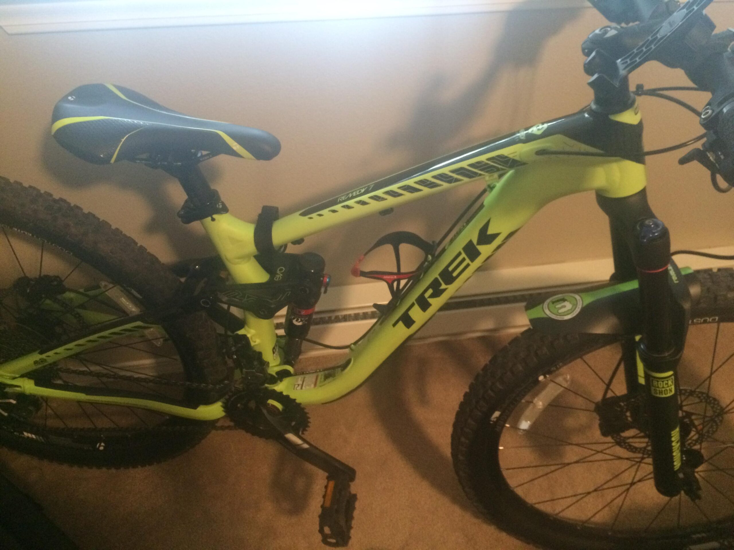 Trek Remedy 7 27.5: A close-up view of a bright green and black Trek mountain bike, featuring a sleek design and a padded seat. The bicycle is positioned indoors on a carpet, with visible details such as the handlebars, brakes, and gearing system. The wheels have a rugged tread pattern suitable for off-road cycling.