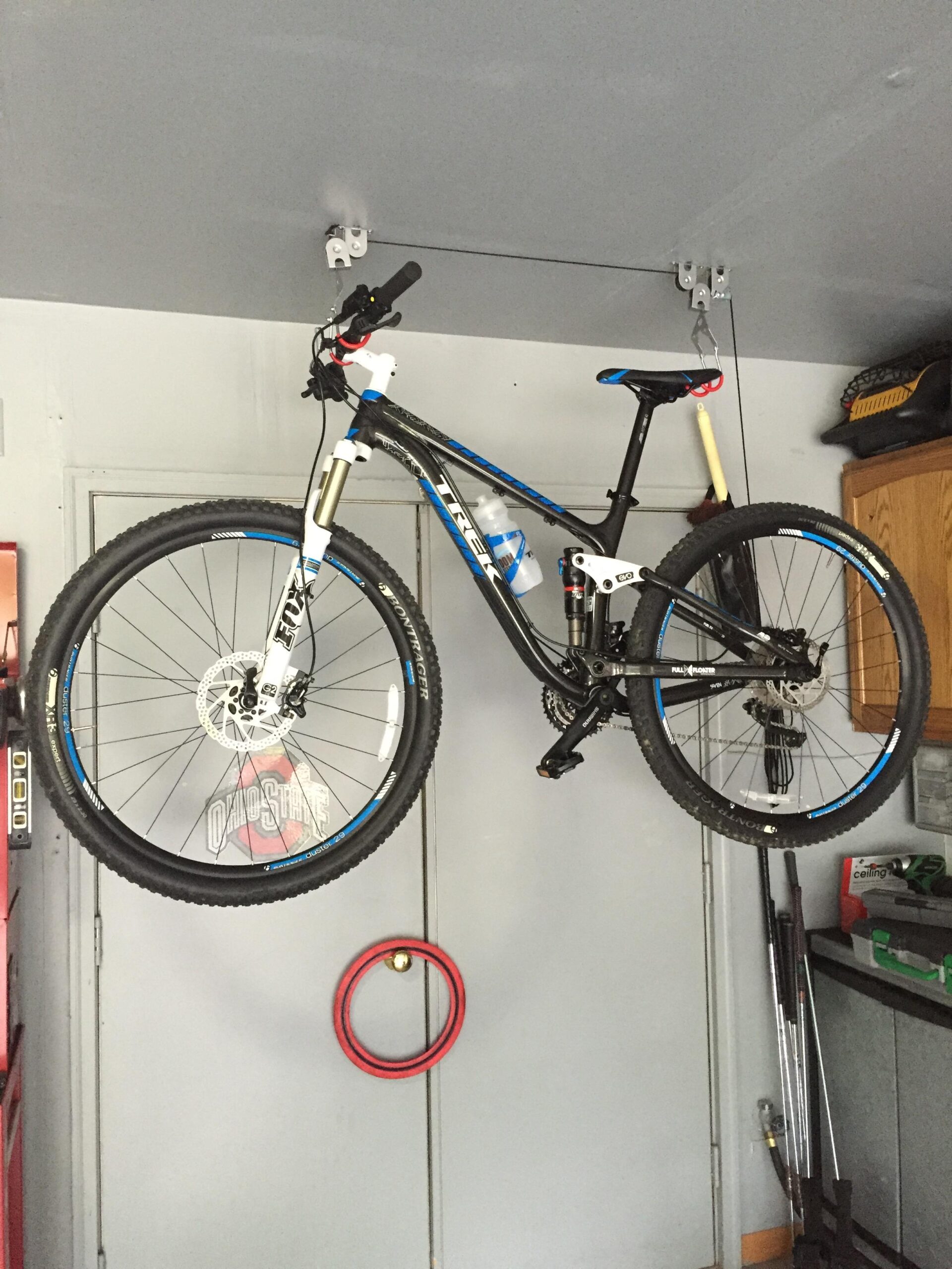 Trek Fuel EX 7 29: A mountain bike hanging from a ceiling-mounted storage system in a garage, with a gray wall and various tools and equipment visible in the background.