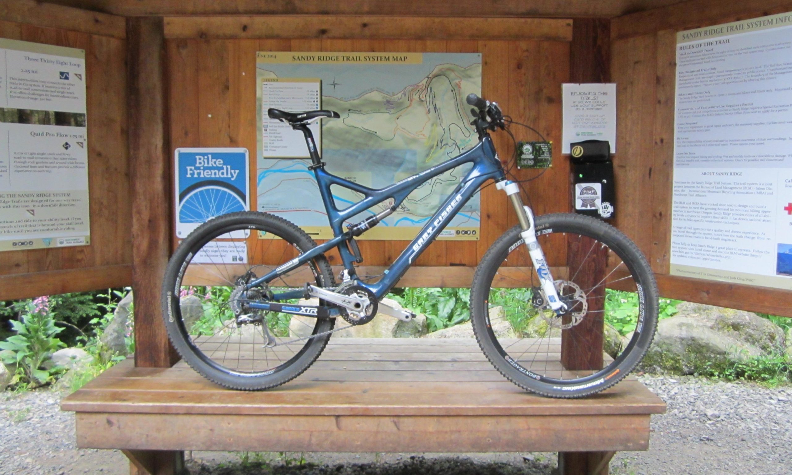 A blue mountain bike is leaning against a wooden information kiosk that displays a trail map and various biking-related signage. The kiosk is surrounded by greenery, indicating an outdoor environment suitable for biking. Various trails and rules are detailed on the signage attached to the walls of the kiosk. Sandy Ridge mountain bike trail.