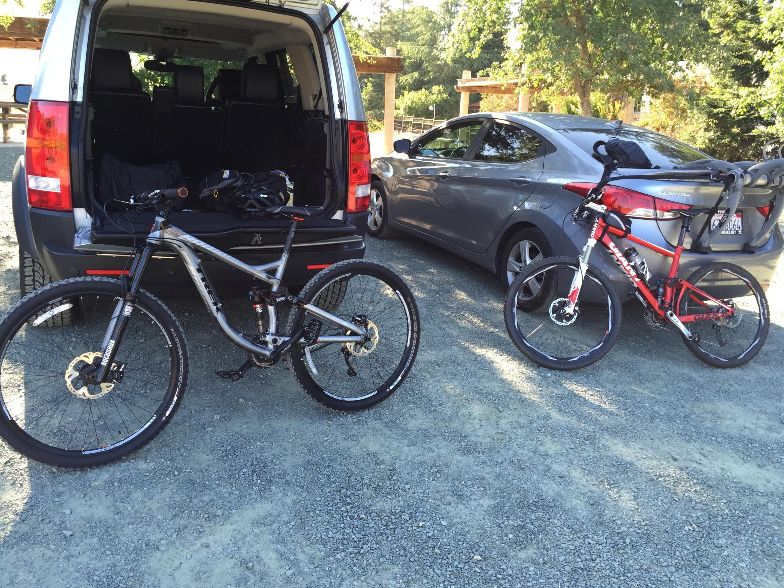 Trek Remedy 7 29: Two bicycles are parked on a gravel surface next to a car and an open trunk of an SUV. One bike is silver with thick tires, while the other is red with thinner tires. The SUV is black with spacious seating visible, and a gray sedan is parked nearby. Lush greenery and wooden structures are in the background, indicating an outdoor setting.