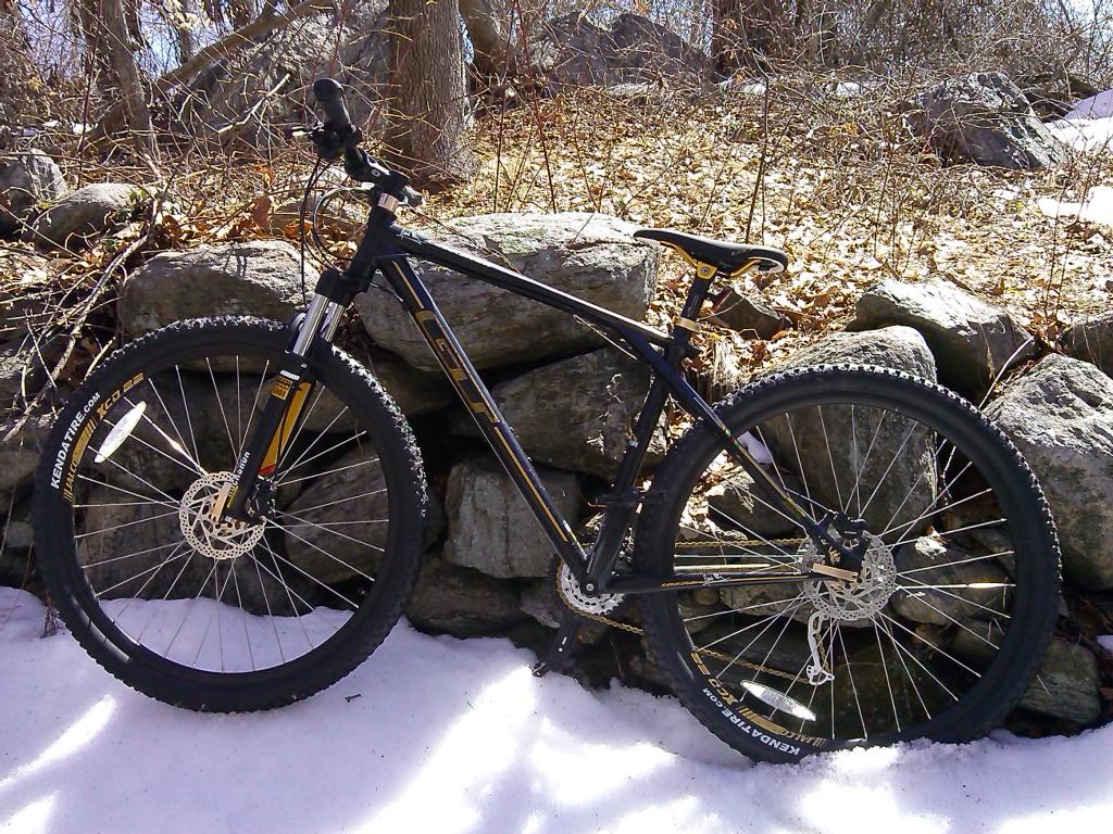 GT Karakoram Elite 2014: A mountain bike leaned against a stone wall in a snowy landscape, with patches of exposed ground and dry leaves in the background. The bike features thick tires, a black and yellow color scheme, and is positioned on a bed of snow.