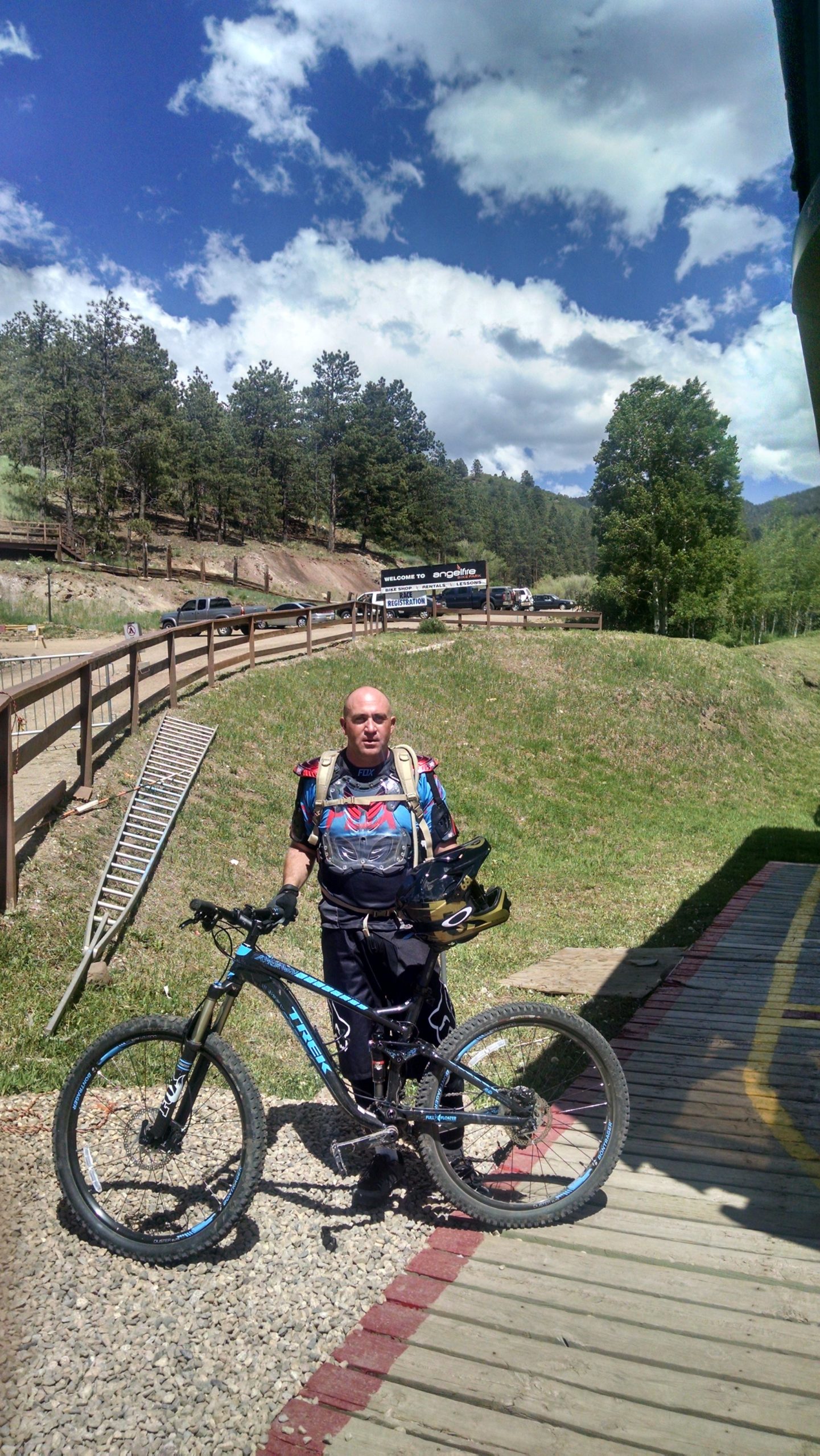 A cyclist in protective gear stands beside a mountain bike at a trailhead. In the background, there are trees and a blue sky with fluffy clouds, along with a sign welcoming visitors to a mountain resort. The cyclist is holding a helmet and appears ready for a ride. Angel Fire Bike Park mountain bike trail.