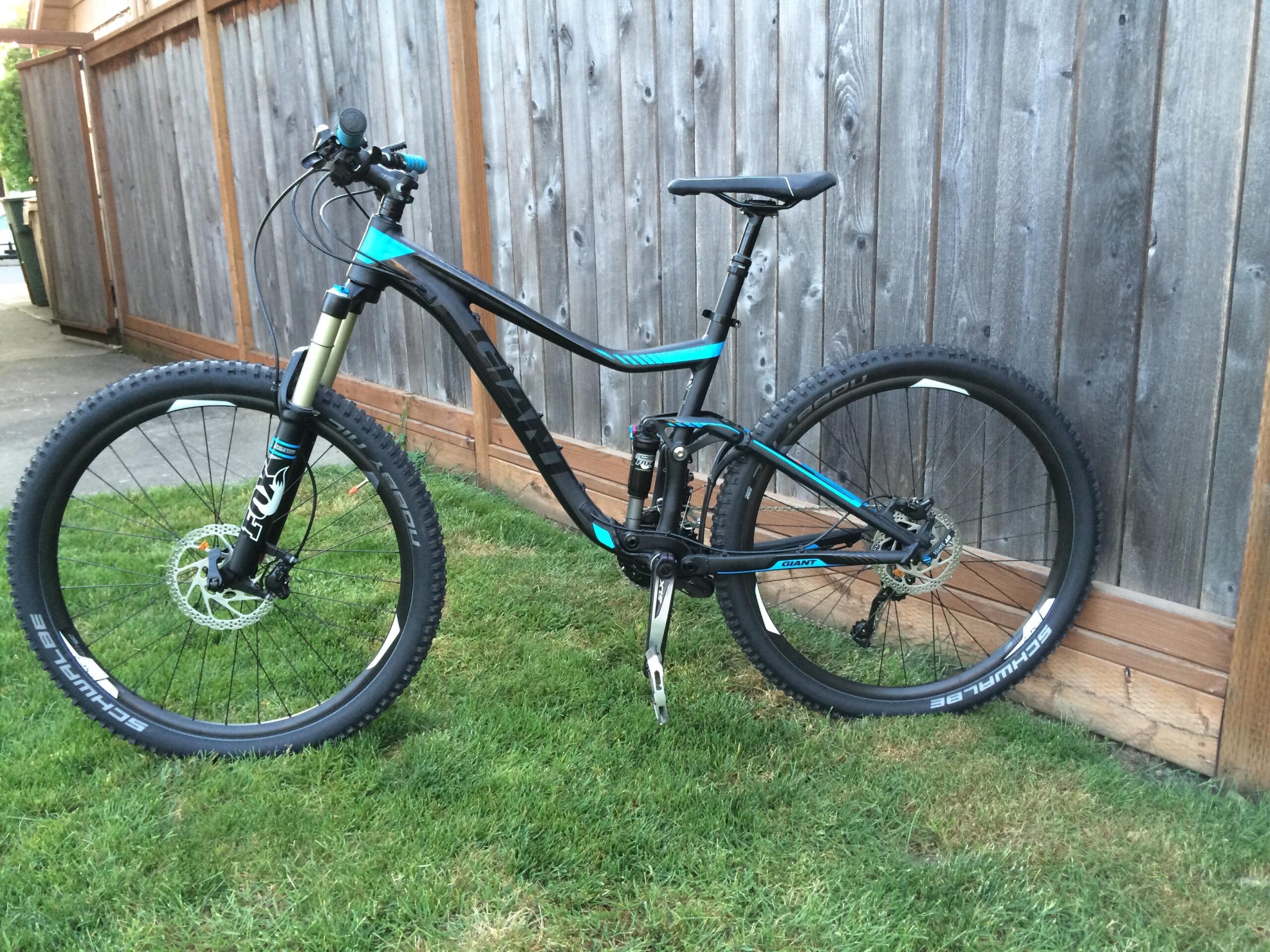 Giant Trance 27.5 2: A black and teal mountain bike is leaning against a wooden fence, resting on a patch of grass. The bike features thick tires, a front suspension fork, and a sleek frame design, showcasing its suitability for off-road riding.