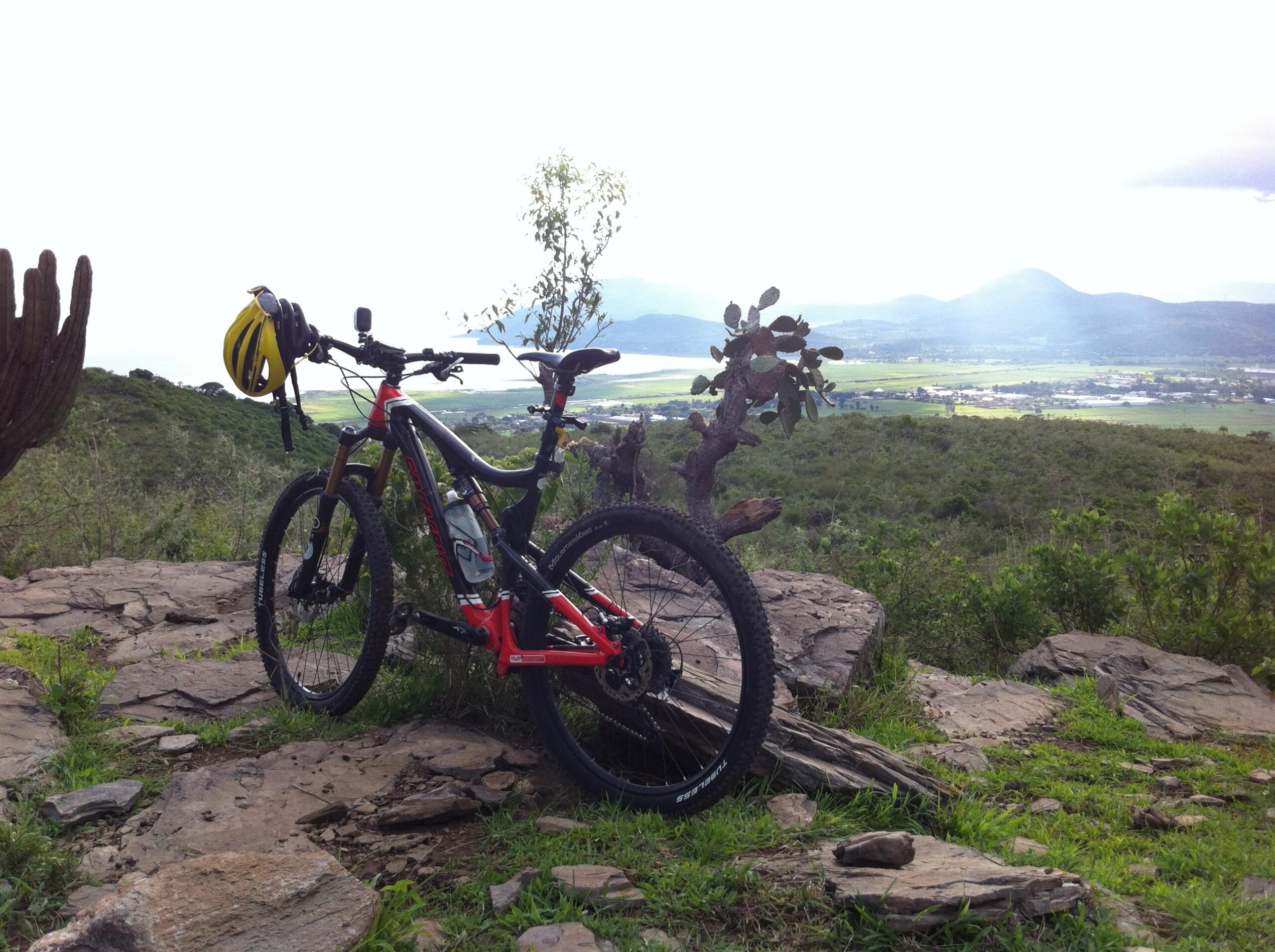 Santa Cruz Blur TRC: A mountain bike with a yellow helmet resting on the handlebars stands on rocky terrain, surrounded by lush greenery. In the background, a scenic view of hills and a valley stretches out under a cloudy sky.
