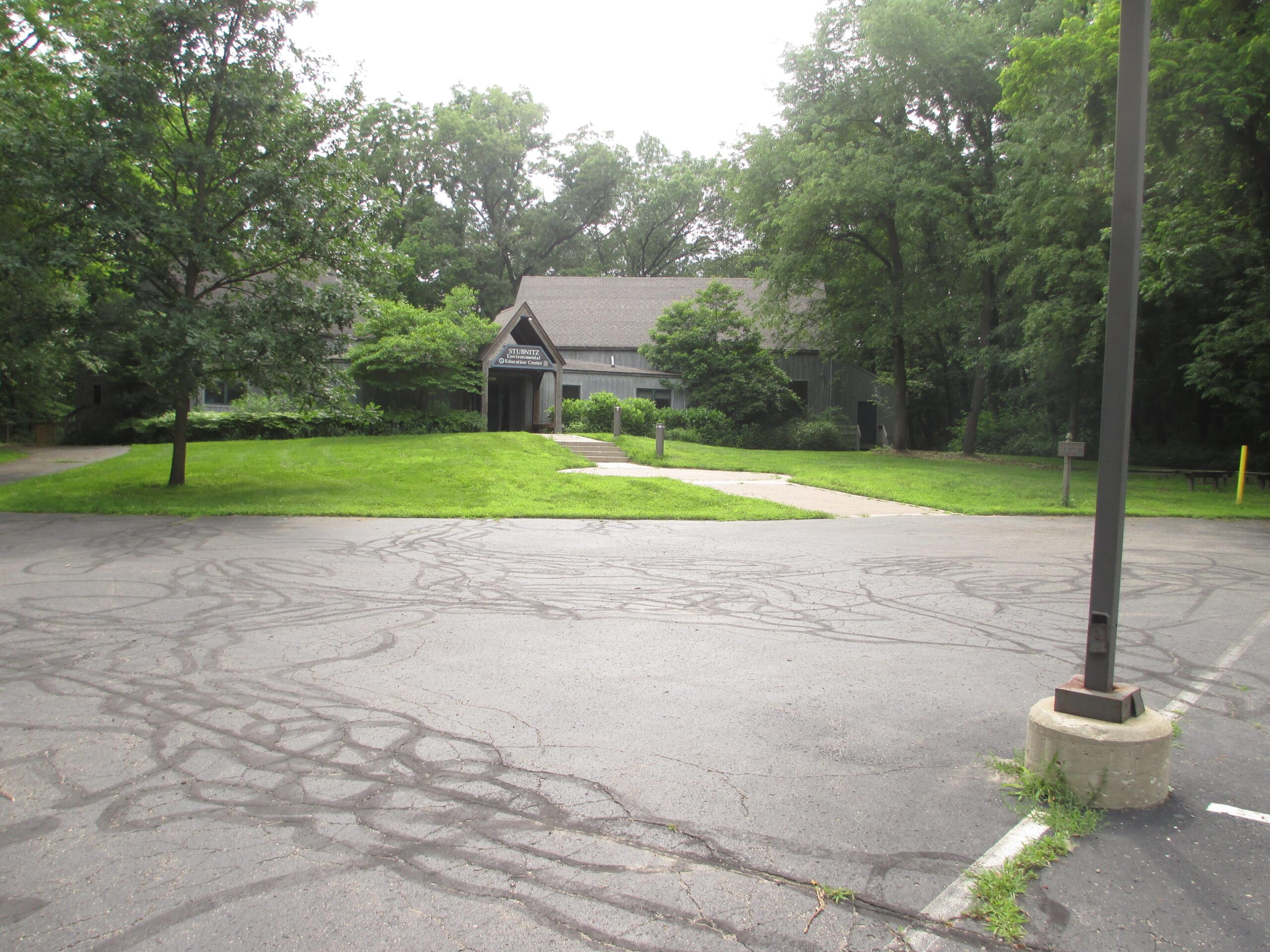 A view of a grassy area leading to a large, brown building surrounded by trees. The foreground features a paved parking lot with tire marks, and a light pole is visible on the right side of the image. The scene is set in a mostly overcast environment with lush greenery. Heritage Park mountain bike trail.