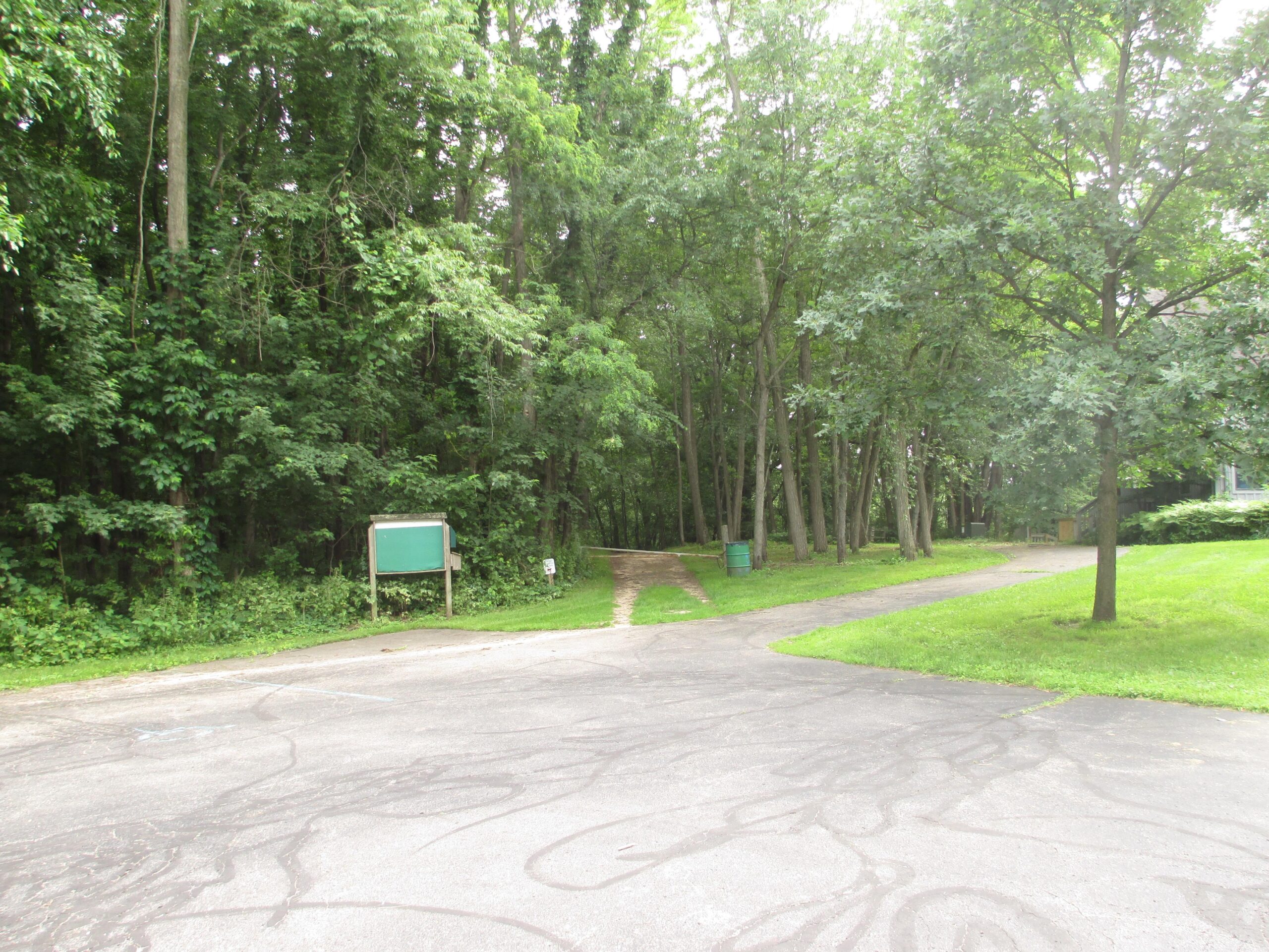 A pathway leading into a dense forest, with a green information board on the left and grassy areas surrounding the paved walkway. The scene is lush and green, indicating a natural outdoor setting. Heritage Park mountain bike trail.
