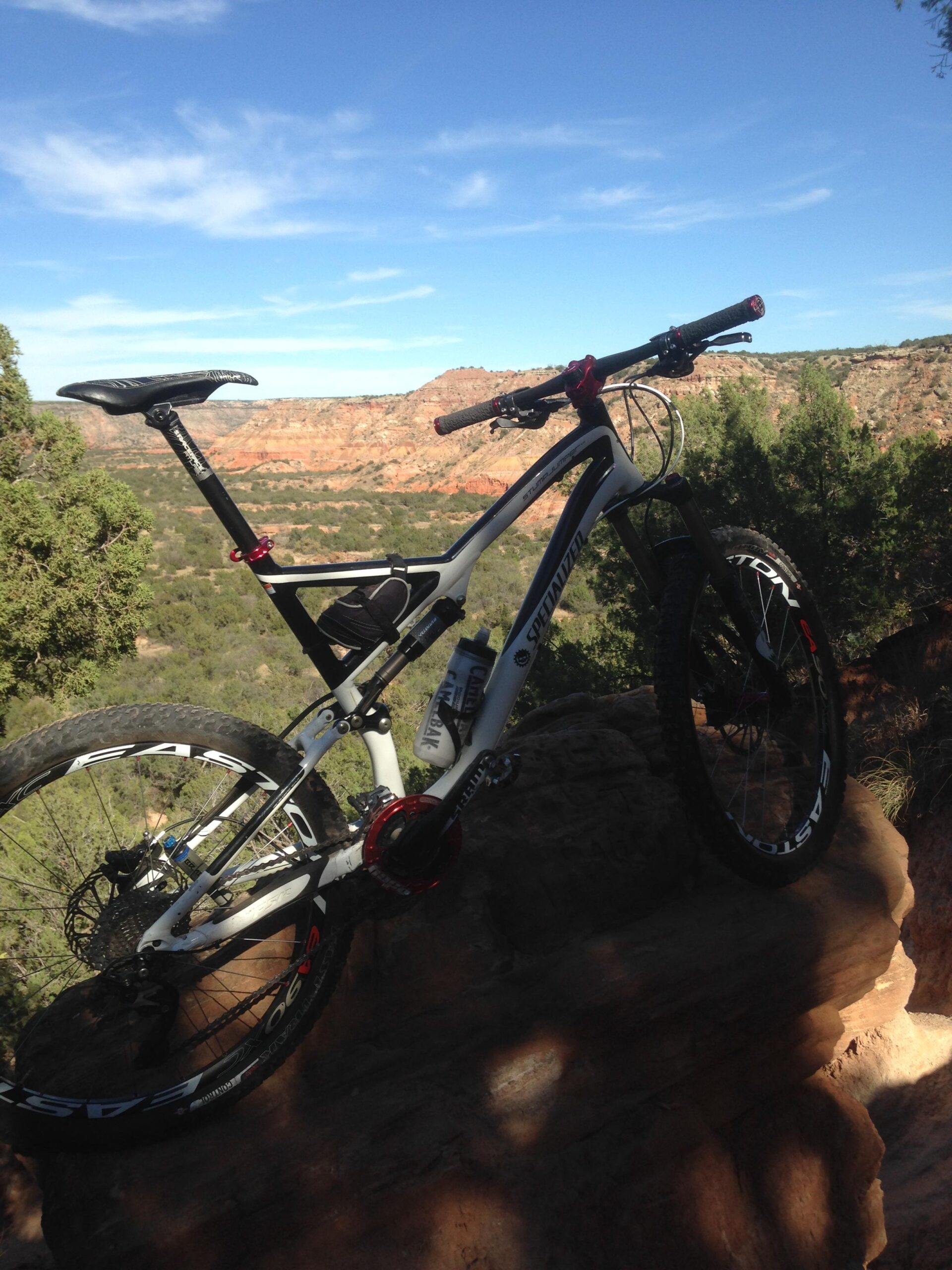Specialized Stumpjumper FSR Elite: A mountain bike positioned on a rocky outcrop, overlooking a scenic landscape with red rock formations and green foliage under a blue sky with scattered clouds.