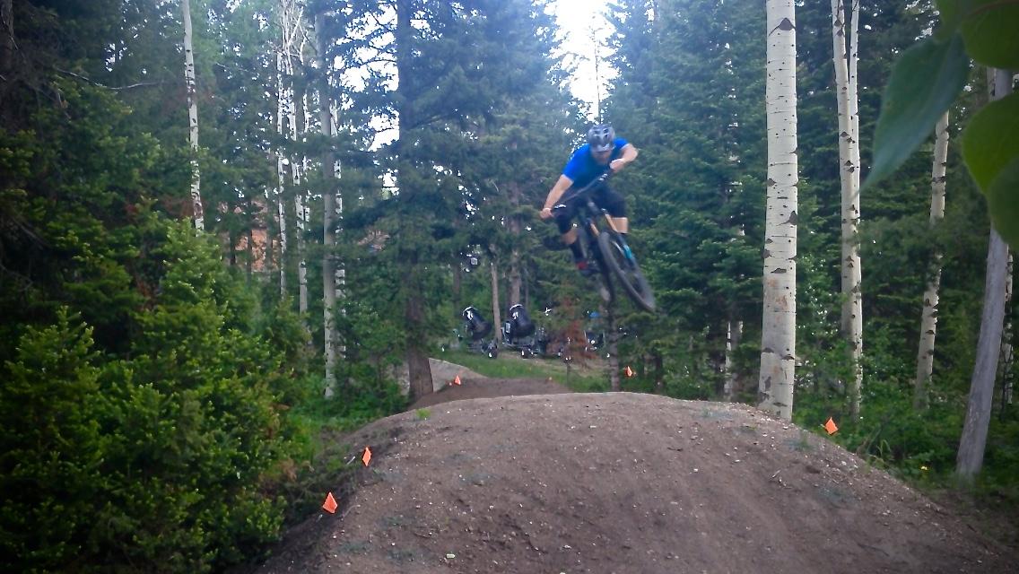 A mountain biker in a blue shirt performs a jump over a dirt ramp in a forested area, surrounded by tall trees and greenery, with orange flags marking the trail. Jackson Hole Bike Park mountain bike trail.
