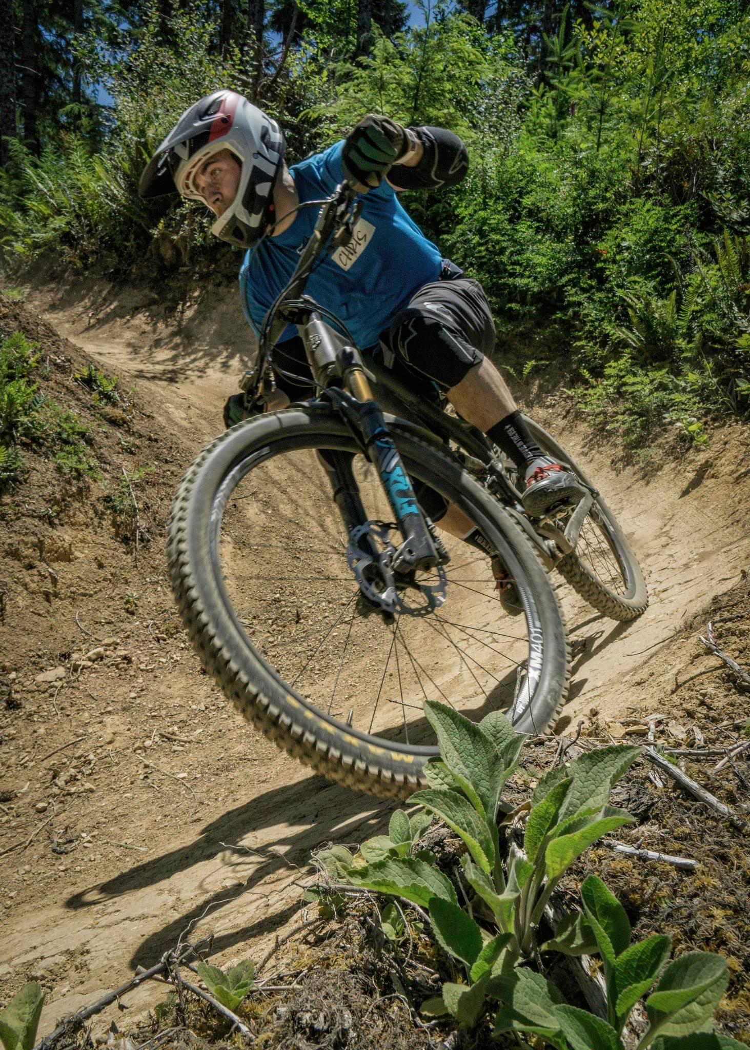 A mountain biker leans into a turn on a dirt trail, surrounded by greenery. The rider is wearing a helmet and protective gear, showcasing dynamic motion as they navigate the path. The image captures the thrill of mountain biking in a natural environment. Alsea Falls Recreation Site mountain bike trail.