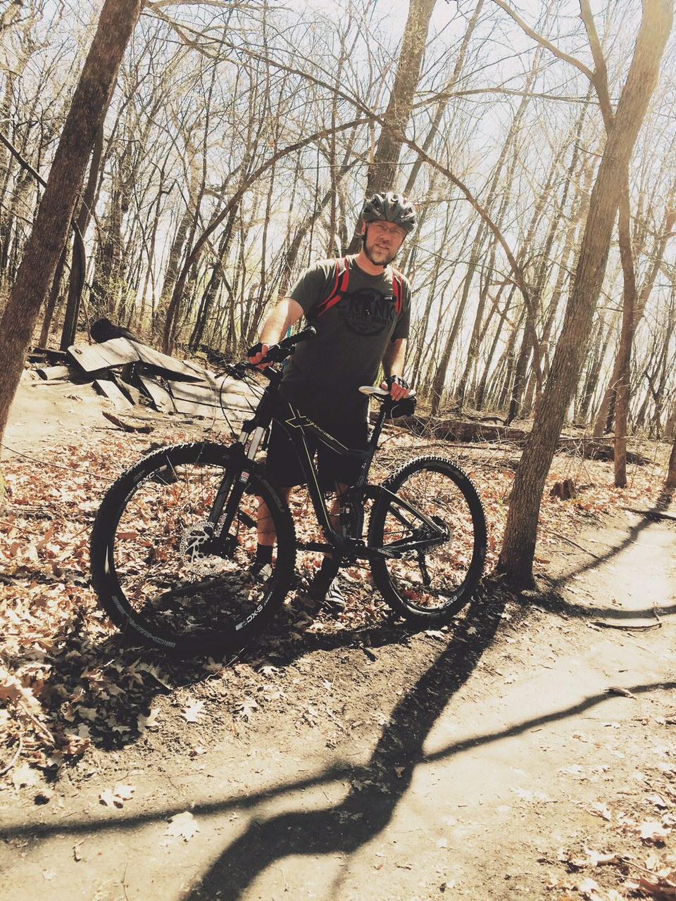 A person standing next to a mountain bike on a wooded trail with bare trees and fallen leaves. The individual is wearing a helmet and a t-shirt, and has a backpack on. Sunlight filters through the trees, casting shadows on the ground. Elm Creek Park mountain bike trail.