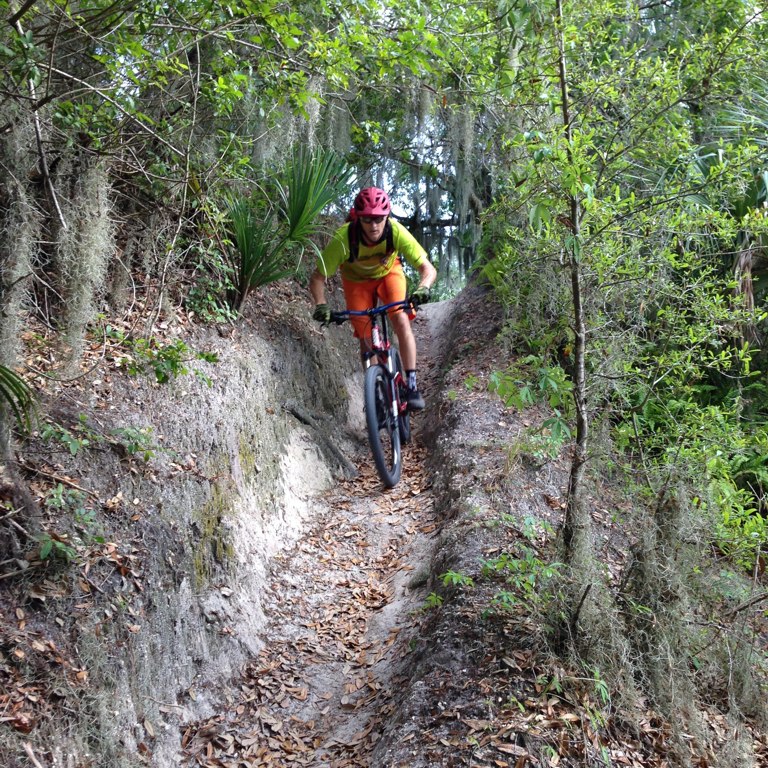 A mountain biker rides down a narrow, dirt trail surrounded by greenery and trees. The biker is wearing a bright yellow shirt, orange shorts, and a red helmet, capturing a moment of action as they navigate the rugged terrain. Leaves and small plants are visible along the sides of the trail, adding to the natural setting. Loyce E. Harpe Park mountain bike trail.