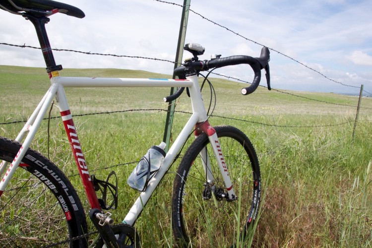 A white and red gravel bike leaning against a barbed wire fence, with a grassy field and rolling hills in the background under a partly cloudy sky. A water bottle is attached to the frame near the pedals, and the bike features black tires and handlebars.
