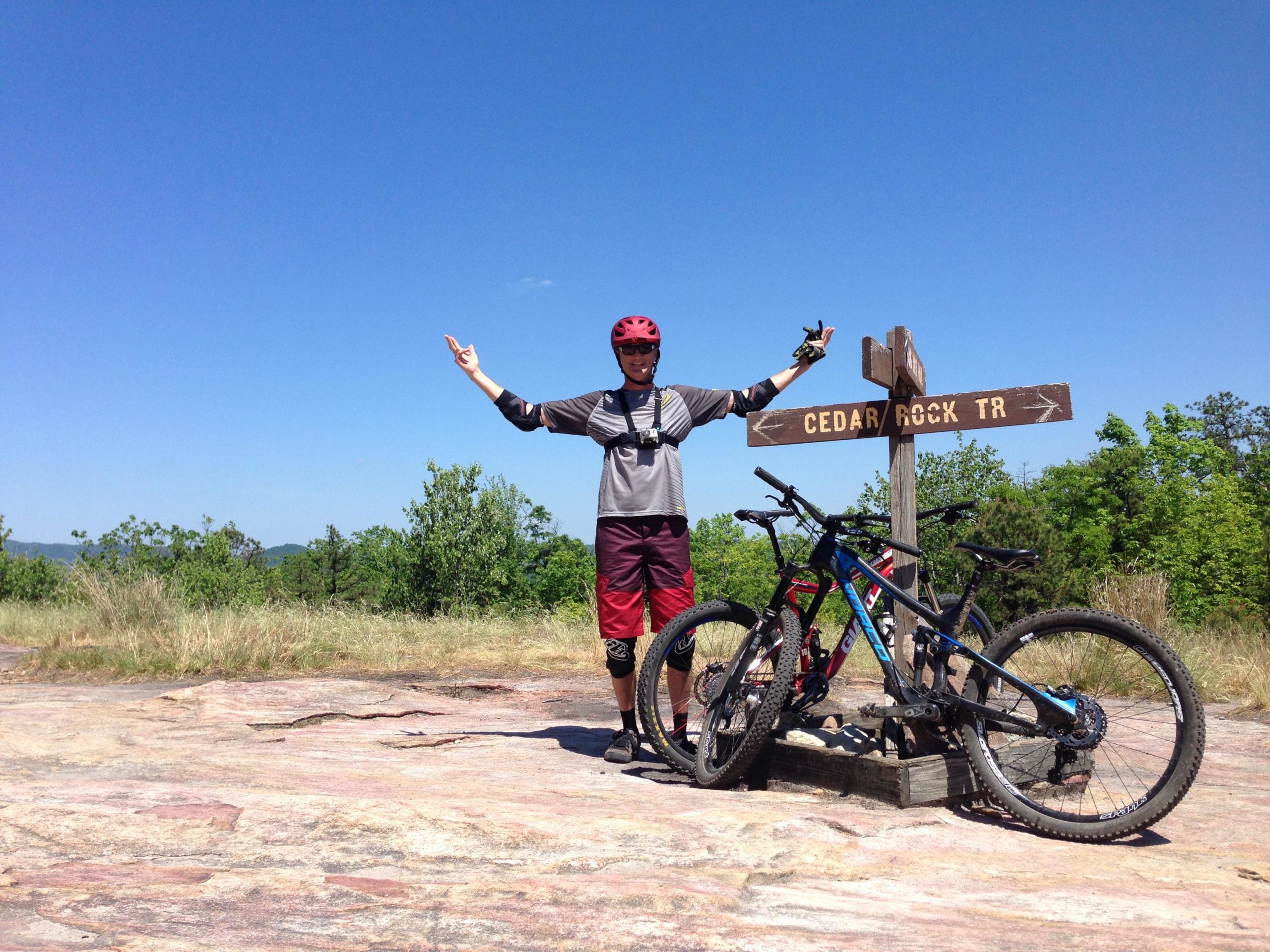 A mountain biker in a helmet and protective gear stands triumphantly next to a wooden signpost marking the Cedar Rock Trail. Two mountain bikes are leaning against the sign, with a clear blue sky and lush green trees in the background. The biker is raising one arm while giving a peace sign with the other, exuding a sense of accomplishment and joy. DuPont State Forest mountain bike trail.