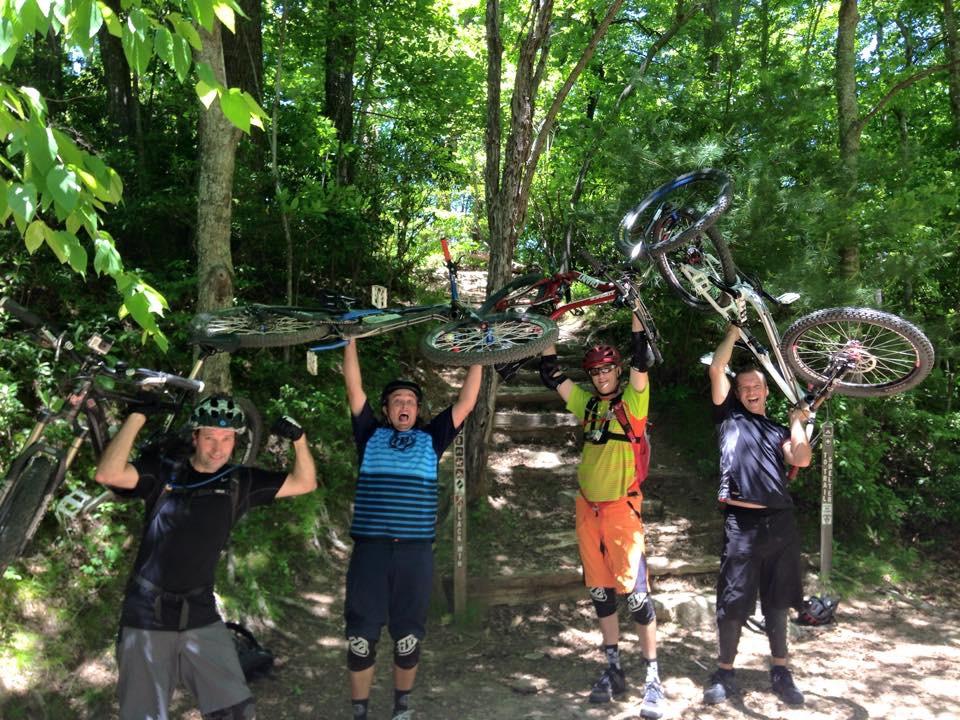 Four mountain bikers pose together outdoors, each lifting their bicycles above their heads in celebration. The group is surrounded by lush greenery and trees, with a dirt path visible behind them. They are wearing cycling gear, including helmets and protective pads, and are smiling enthusiastically. Black Mountain mountain bike trail.
