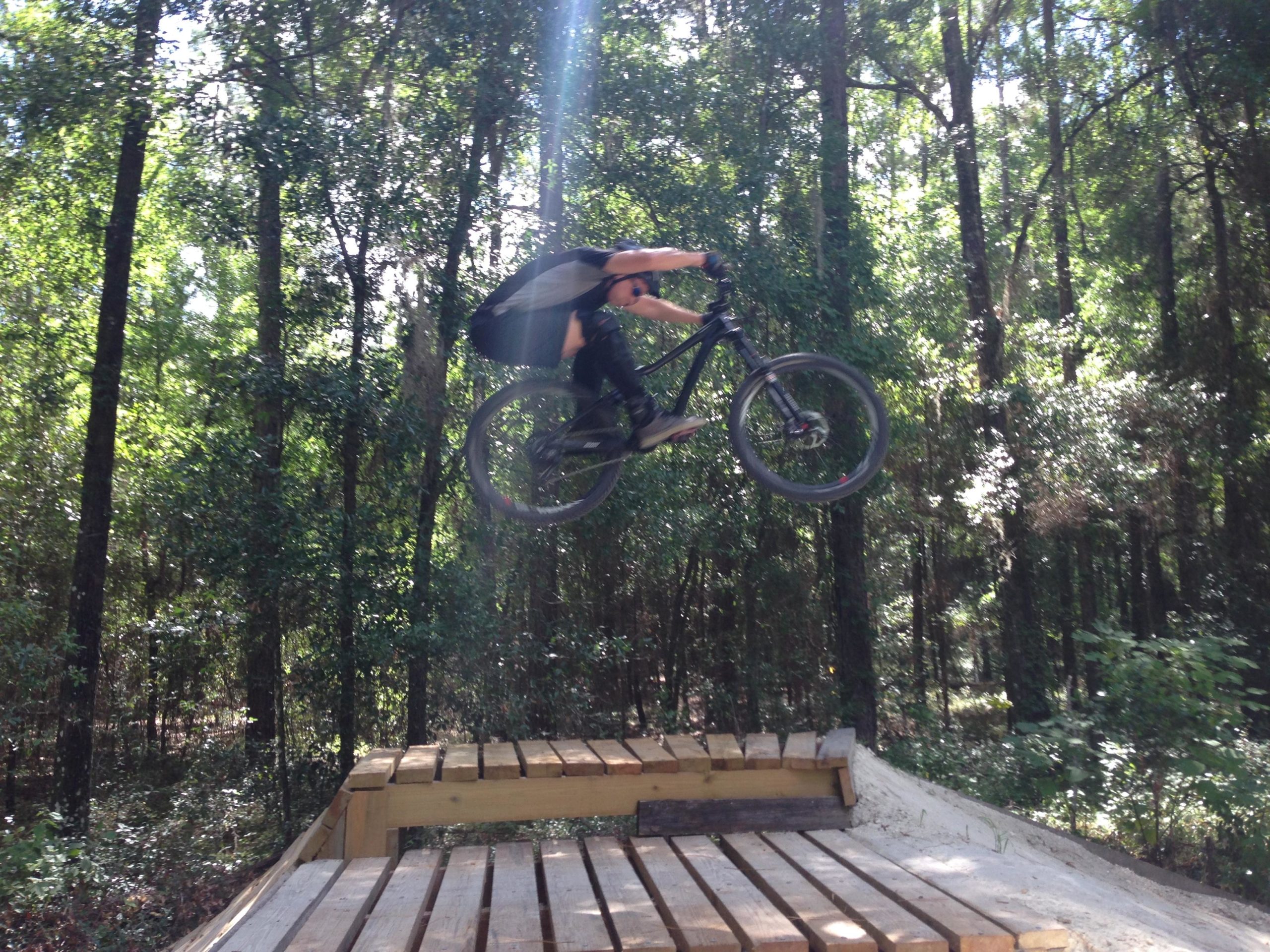 A person riding a mountain bike performs a jump off a wooden ramp in a dense forest setting, with sunlight streaming through the trees. Santos mountain bike trail.