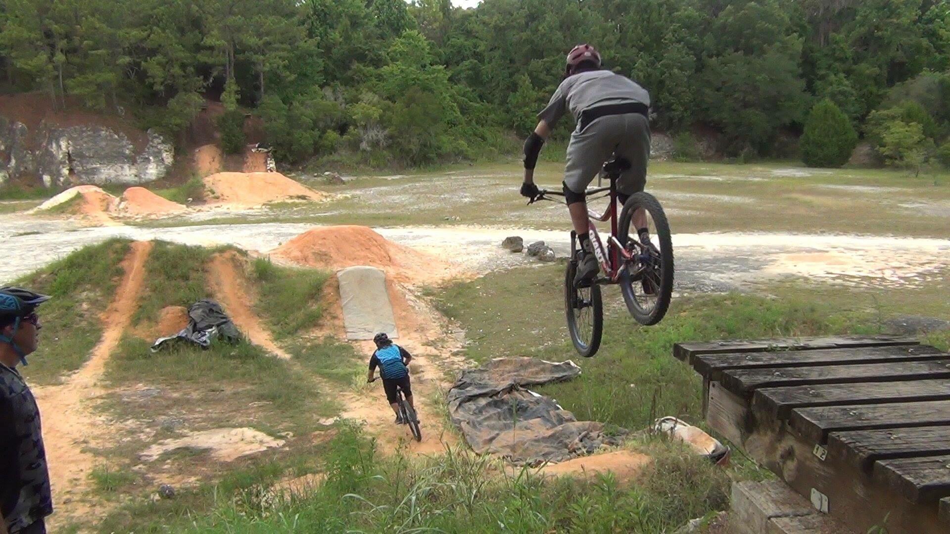 Two mountain bikers are seen in a dirt bike park: one rider launching off a ramp in the air, while the other rides down a dirt trail. The background features dirt jumps and greenery. Santos mountain bike trail.