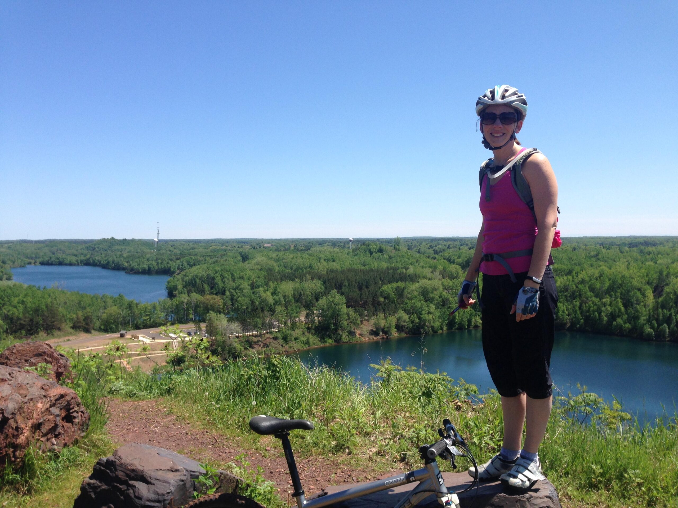 A cyclist standing on a rocky overlook, wearing a pink tank top, helmet, and gloves, smiling at the camera. In the background, lush greenery and a lake stretch out under a clear blue sky, with scenic hills visible in the distance. A bicycle rests on the ground nearby. Cuyuna Lakes mountain bike trail.