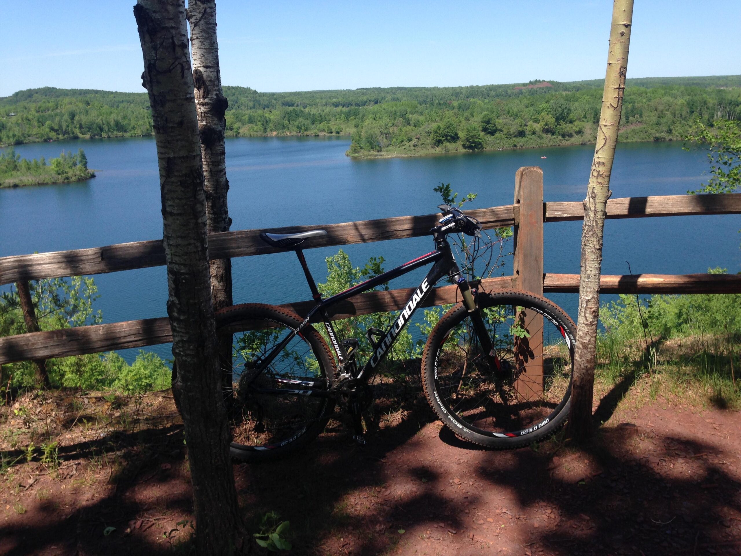 A mountain bike rests against a wooden fence overlooking a serene blue lake surrounded by green trees and hills under a clear blue sky. Cuyuna Lakes mountain bike trail.