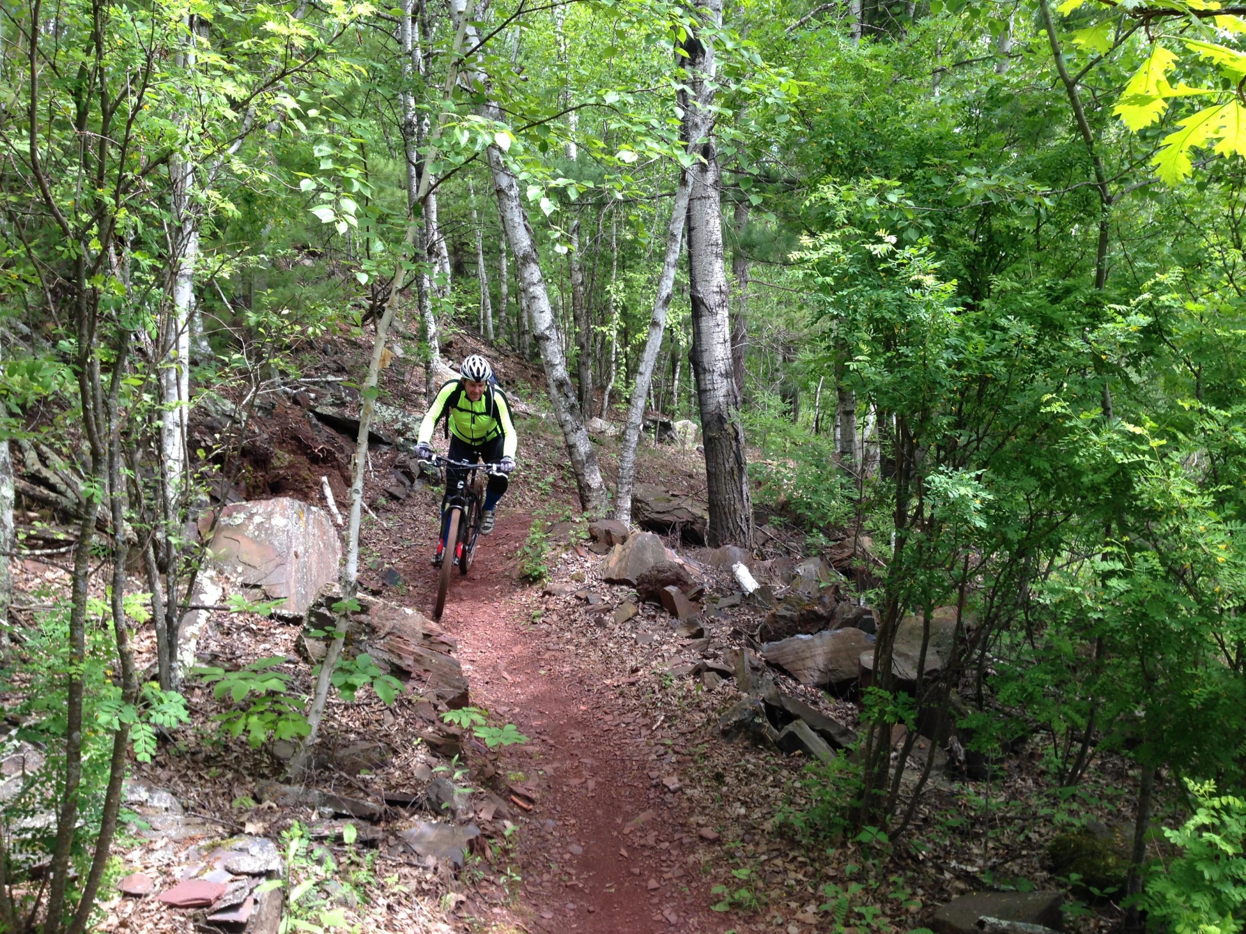 A mountain biker navigates a narrow dirt trail surrounded by lush greenery and rocky terrain in a forested area. The cyclist, wearing a helmet and bright gear, leans slightly forward as they ride through the scenic landscape. Cuyuna Lakes mountain bike trail.