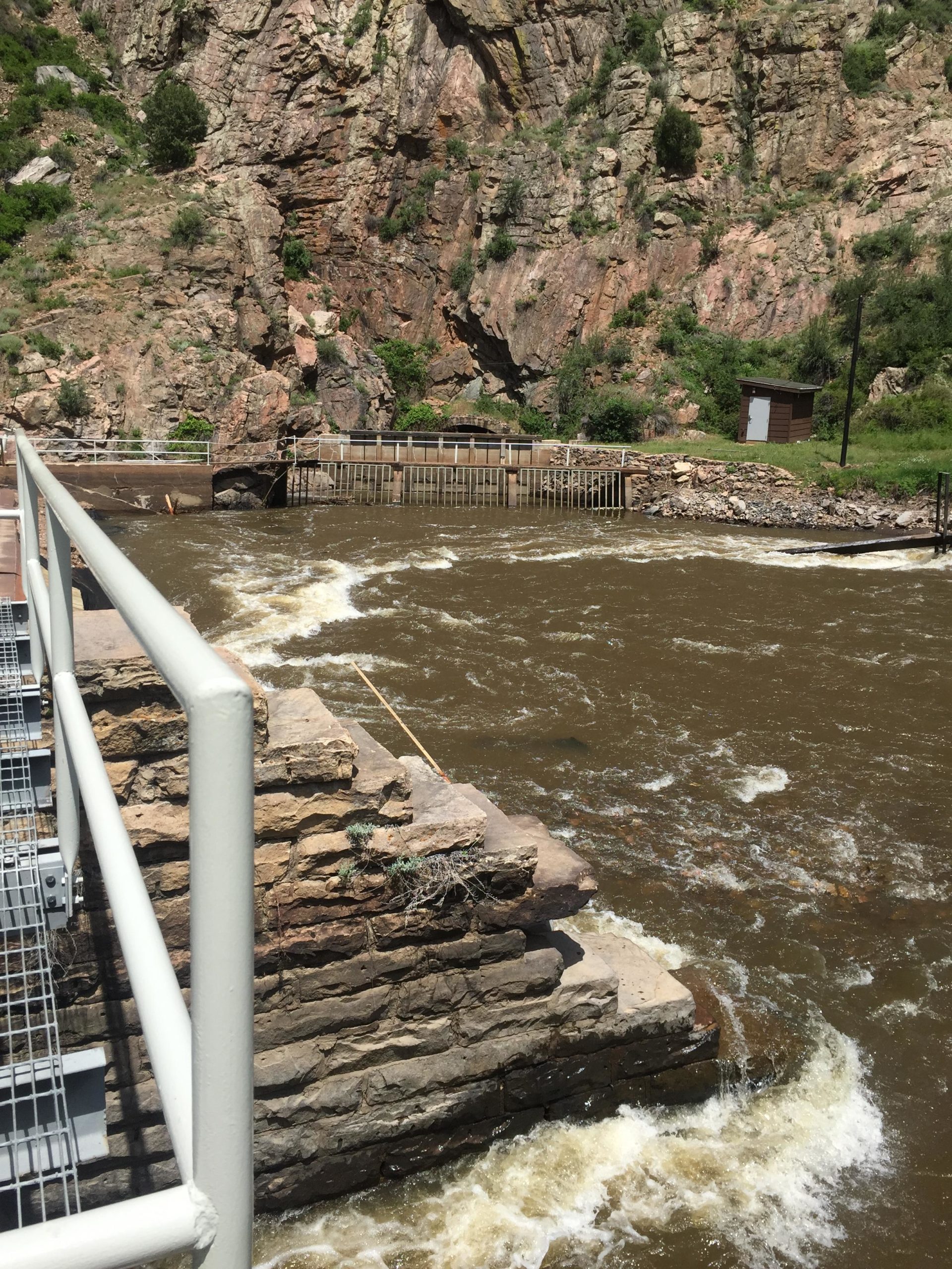 A view of a river flowing past rocky banks, with a wooden structure visible in the background. The foreground features a stone ledge and a railing, while lush greenery and rocky cliffs rise on either side of the river. The water is brown and choppy, indicative of movement and current. Waterton Canyon mountain bike trail.