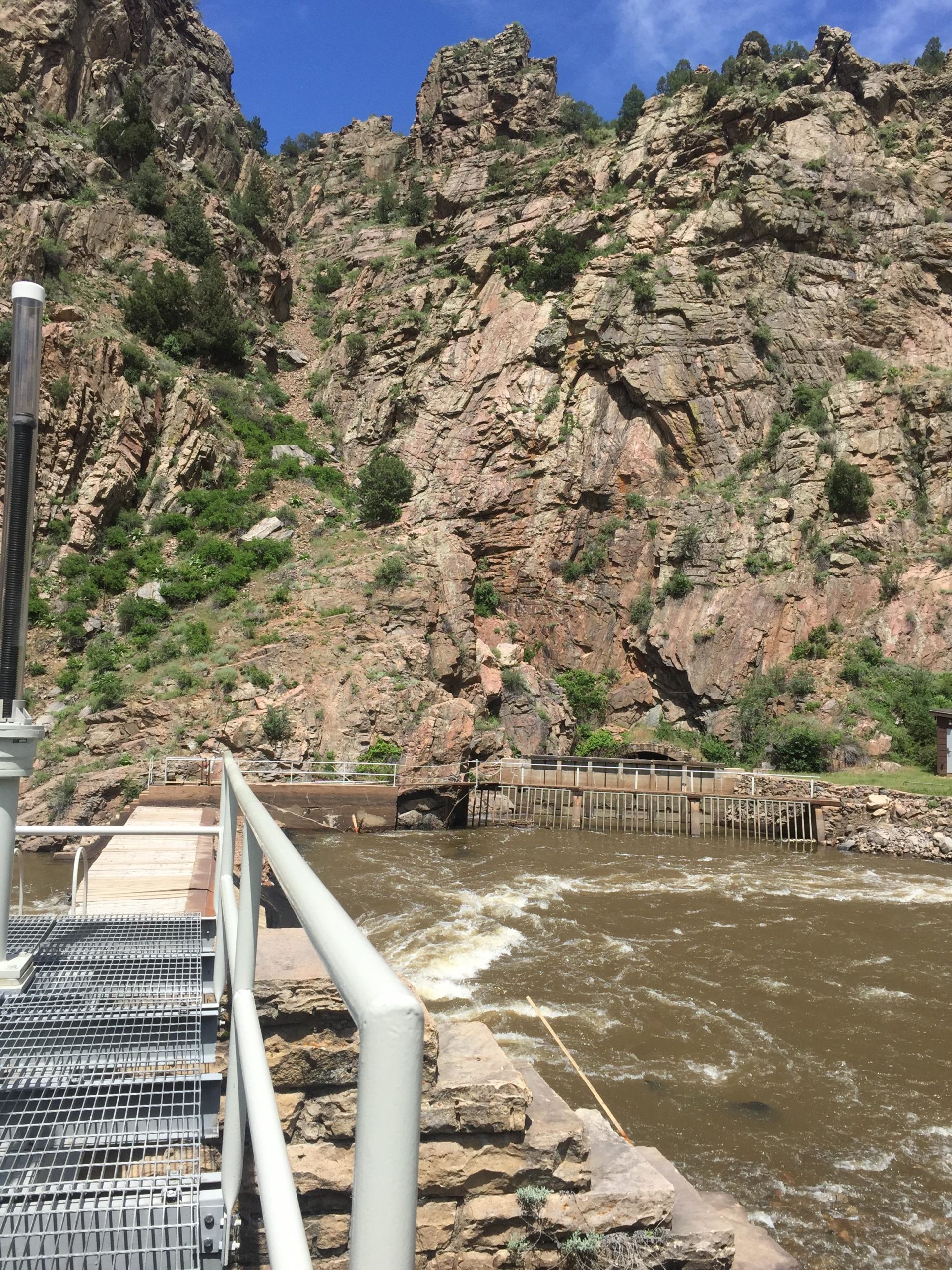 A rocky landscape with steep cliffs and a flowing river in the foreground. A metal walkway extends over the water, leading toward a structure near the riverbank. The scene is set under a blue sky with scattered clouds, showcasing a natural environment with greenery on the slopes. Waterton Canyon mountain bike trail.