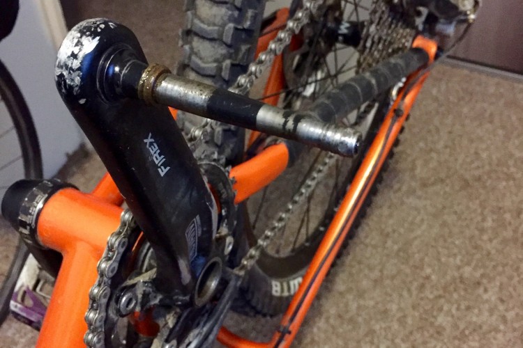 Close-up view of a mountain bike's crankset, showcasing the black crank arm, silver pedal spindle, and orange frame. The bike chain and rear wheel can also be partially seen, indicating regular use and some wear. The background features a carpeted surface.