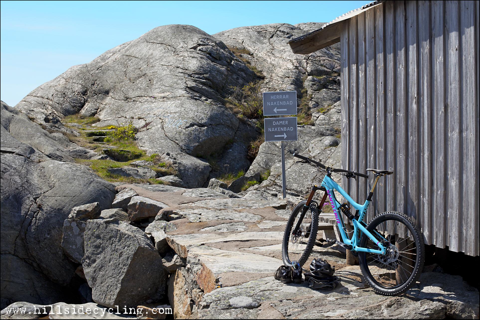 Santa Cruz Nomad Carbon 27.5: A mountain bike rests beside a wooden shed on rocky terrain. Two directional signs indicate paths for men and women to a nearby nudist beach. The background features large, textured rocks and a clear blue sky, emphasizing a serene outdoor setting.