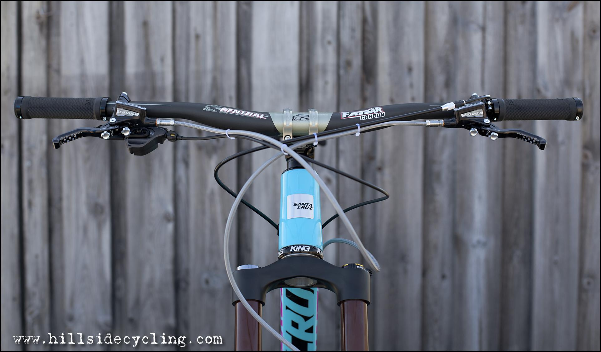 Santa Cruz Nomad Carbon 27.5: Alt text: Close-up view of a mountain bike’s handlebars and stem, featuring black grips, brake levers, and cable routing, set against a weathered wooden background.
