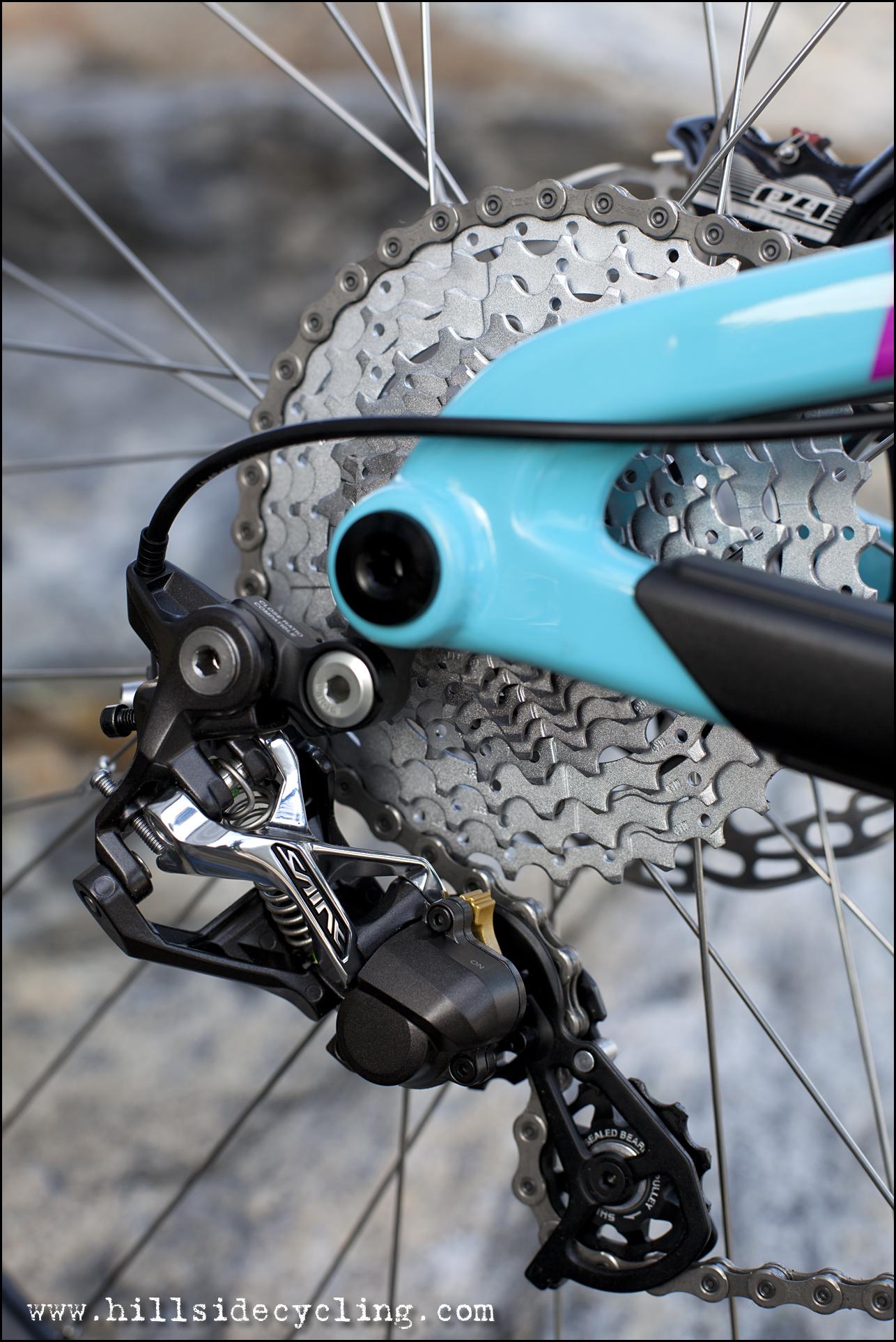Santa Cruz Nomad Carbon 27.5: Close-up of a bicycle's rear derailleur and cassette, showcasing the intricate gears and a turquoise bike frame. The image highlights the mechanical components and chain, set against a blurred background.