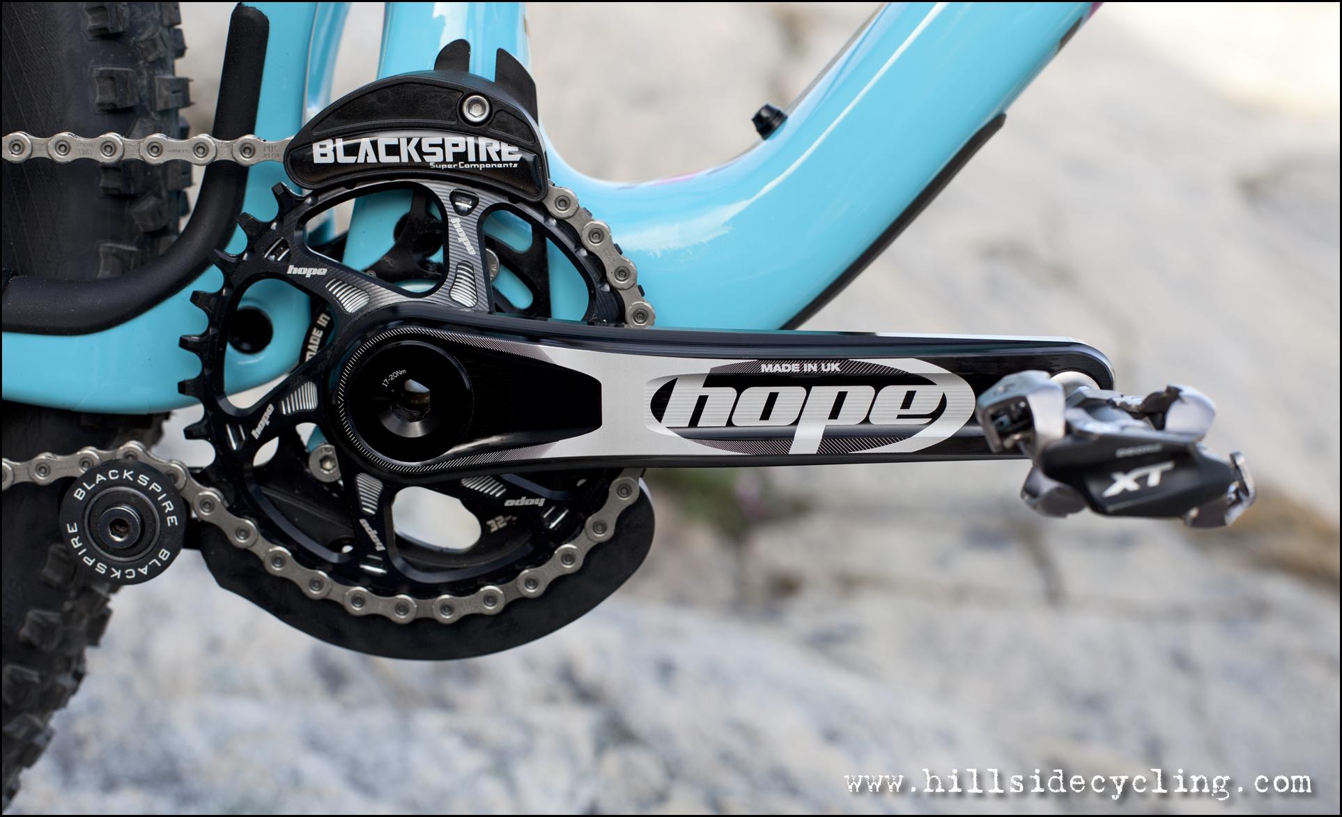 Santa Cruz Nomad Carbon 27.5: Close-up view of a bicycle's drivetrain, featuring a black and silver crankset with "Hope" branding, a black chainring, and a "Blackspire" chain guide. The image shows the bike frame in a light blue color and highlights the rear derailleur and chain, set against a natural rocky background.