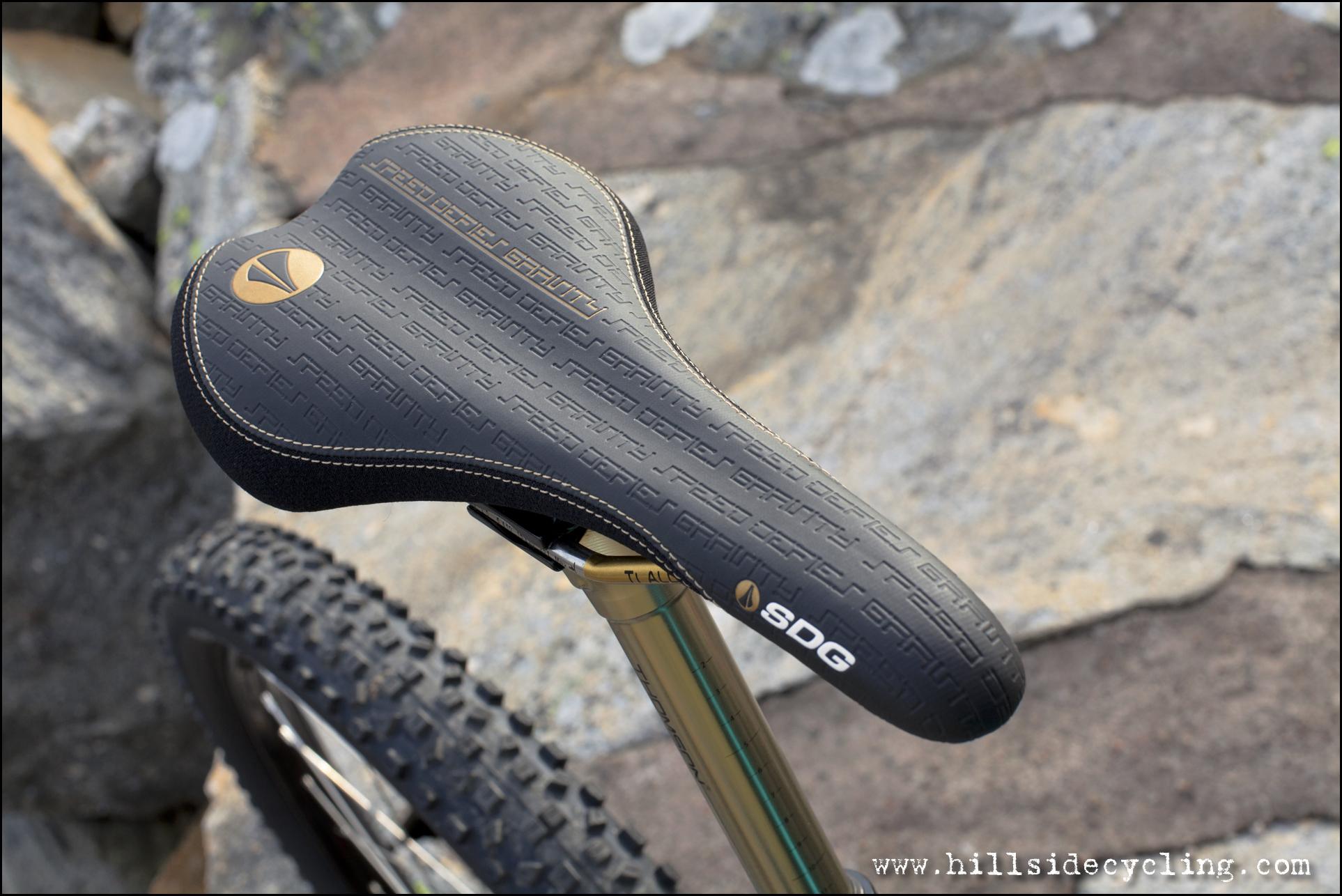 Santa Cruz Nomad Carbon 27.5: Close-up view of a bicycle seat with textured black surface and gold stitching, mounted on a metal seat post. The seat features the branding "SDG" and an embossed design reading "Ready to Ride Gravity." The background shows rocky terrain, suggesting an outdoor cycling environment.