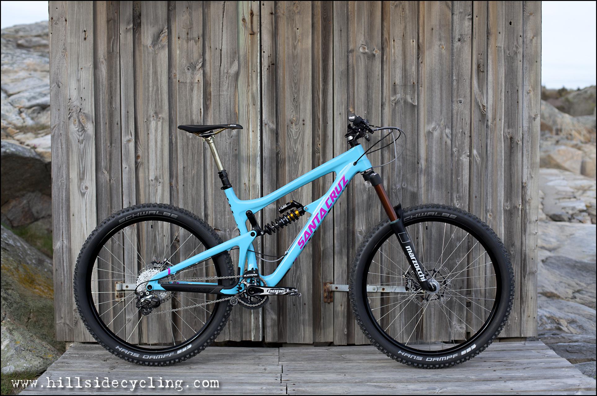 Santa Cruz Nomad Carbon 27.5: A blue mountain bike with a stylish design featuring "Santa Cruz" branding in bold letters is positioned against a wooden backdrop. The bike showcases a modern frame, high-quality suspension components, and knobby tires suitable for off-road trails.
