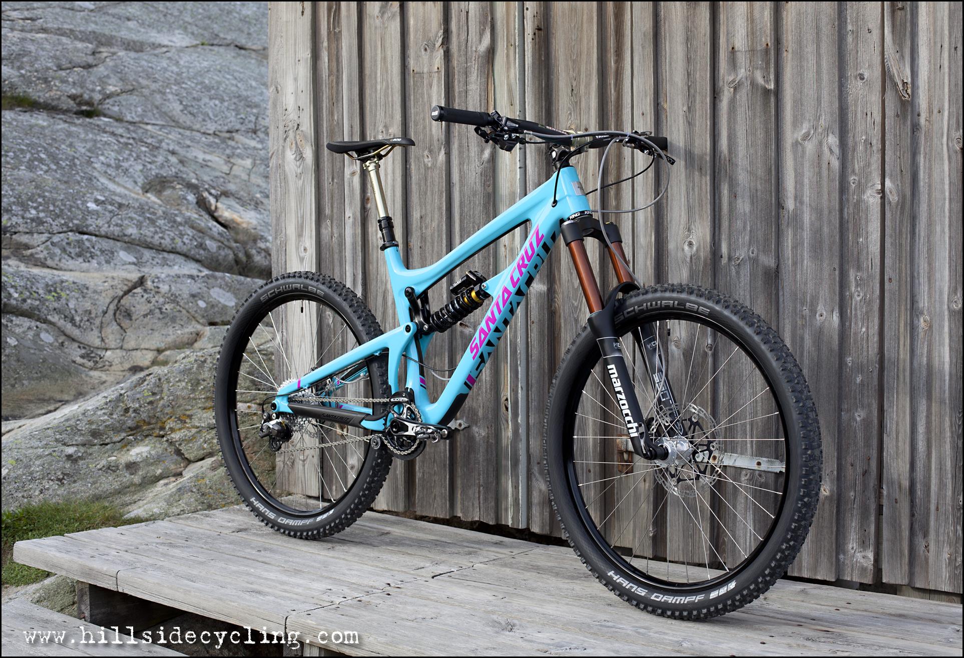 Santa Cruz Nomad Carbon 27.5: A turquoise mountain bike with black wheels and a distinctive design, positioned on a wooden platform in front of a weathered wooden wall. The bike features front suspension forks and a colorful frame labeled "Santa Cruz." The background includes rocky terrain and vegetation, suggesting an outdoor setting suitable for biking.