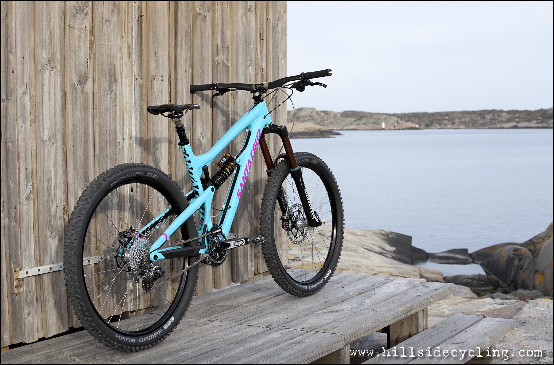 Santa Cruz Nomad Carbon 27.5: A blue and pink mountain bike is leaning against a wooden wall next to a tranquil body of water. The bike features a sleek design with thick tires, a detailed gear system, and a sturdy frame. The background includes a rocky shoreline and distant land formations under a clear sky.