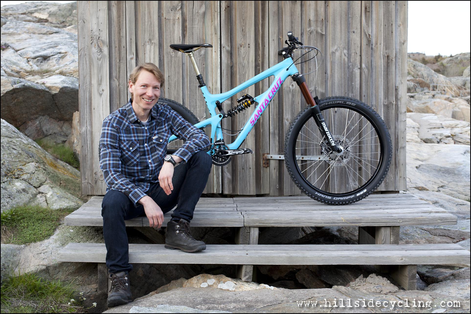 Santa Cruz Nomad Carbon 27.5: A man sitting on wooden steps next to a blue mountain bike with pink accents, smiling at the camera. The background features a rustic wooden wall and rocky terrain. He is dressed in a plaid shirt and dark jeans, with hiking boots visible. The setting appears outdoor and natural.