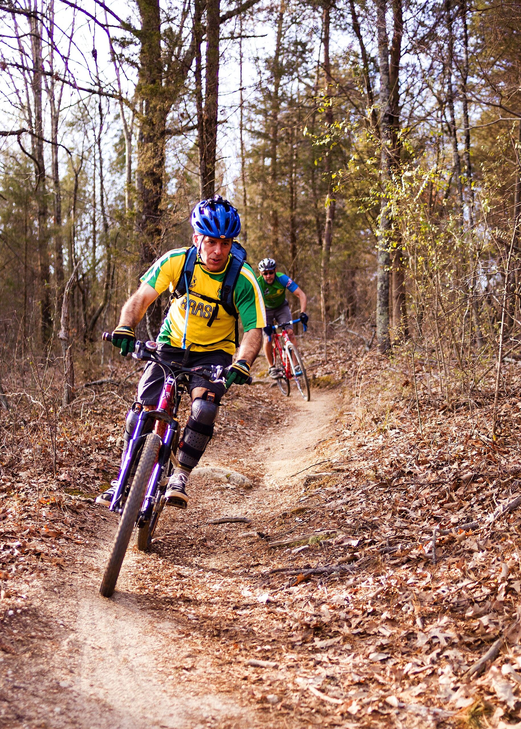 A mountain biker navigating a dirt trail through a wooded area. The rider wears a yellow and green jersey and a blue helmet, demonstrating an advanced skill level as he leans into the turn. A second cyclist follows closely behind, dressed in a green shirt. The ground is covered with fallen leaves and scattered twigs, surrounded by tall trees. Hobby Park mountain bike trail.