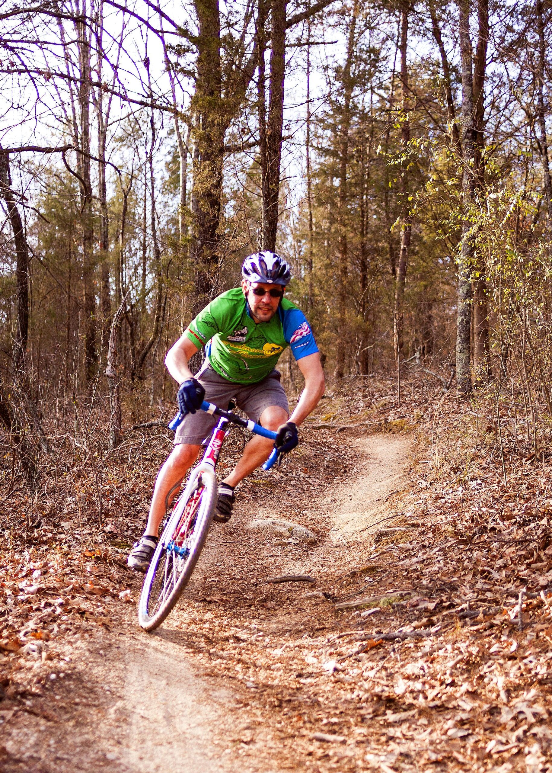 A mountain biker leaning into a turn on a dirt trail surrounded by trees, wearing a green jersey and helmet, with sunglasses on. The ground is covered in fallen leaves, and the trail curves through a forested area. Hobby Park mountain bike trail.
