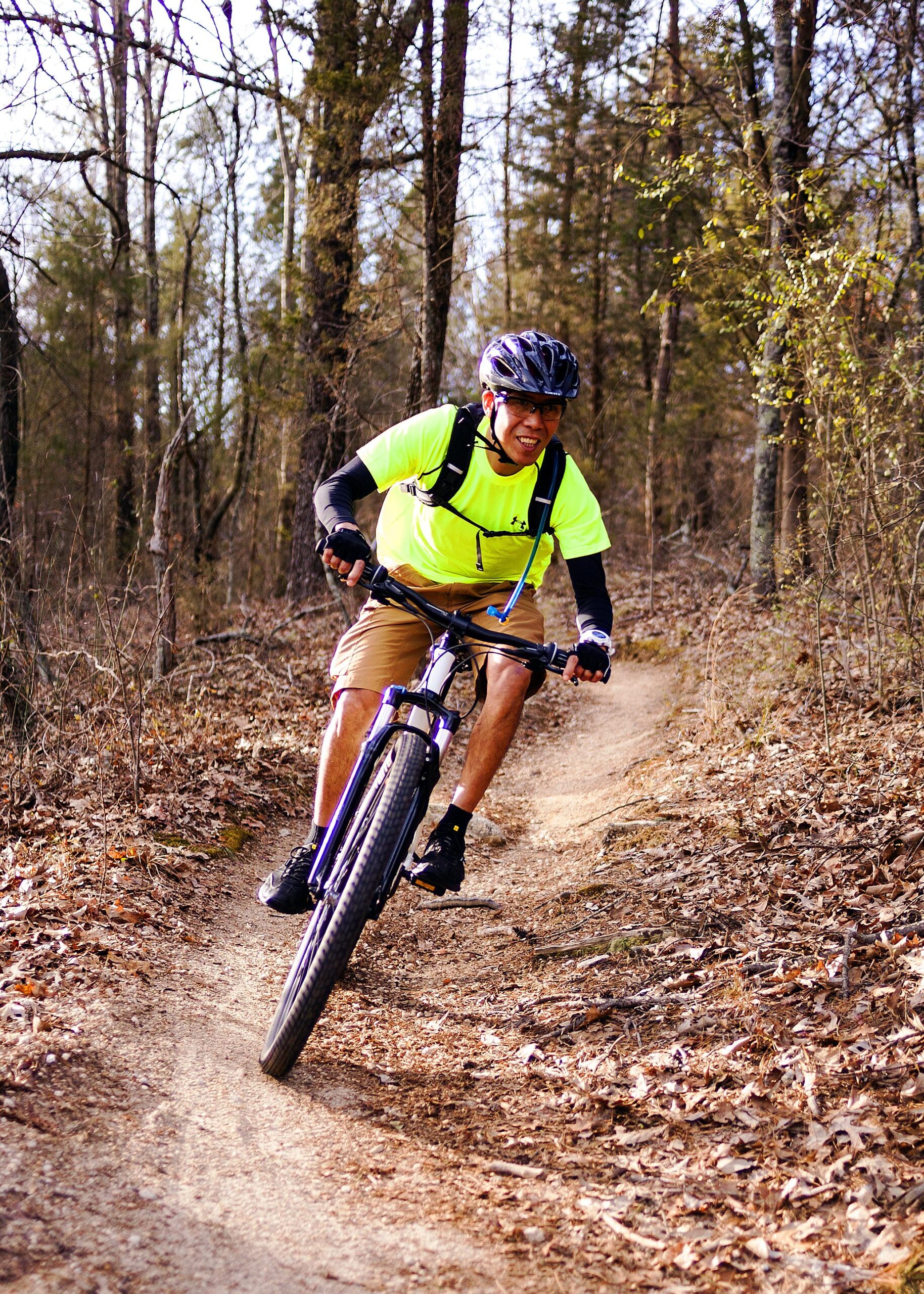 A person wearing a bright yellow shirt and a helmet rides a mountain bike along a winding dirt trail in a wooded area. The surroundings feature trees with sparse leaves, indicative of early spring. The rider has a focused expression as they navigate the trail, with fallen leaves covering the ground. Hobby Park mountain bike trail.