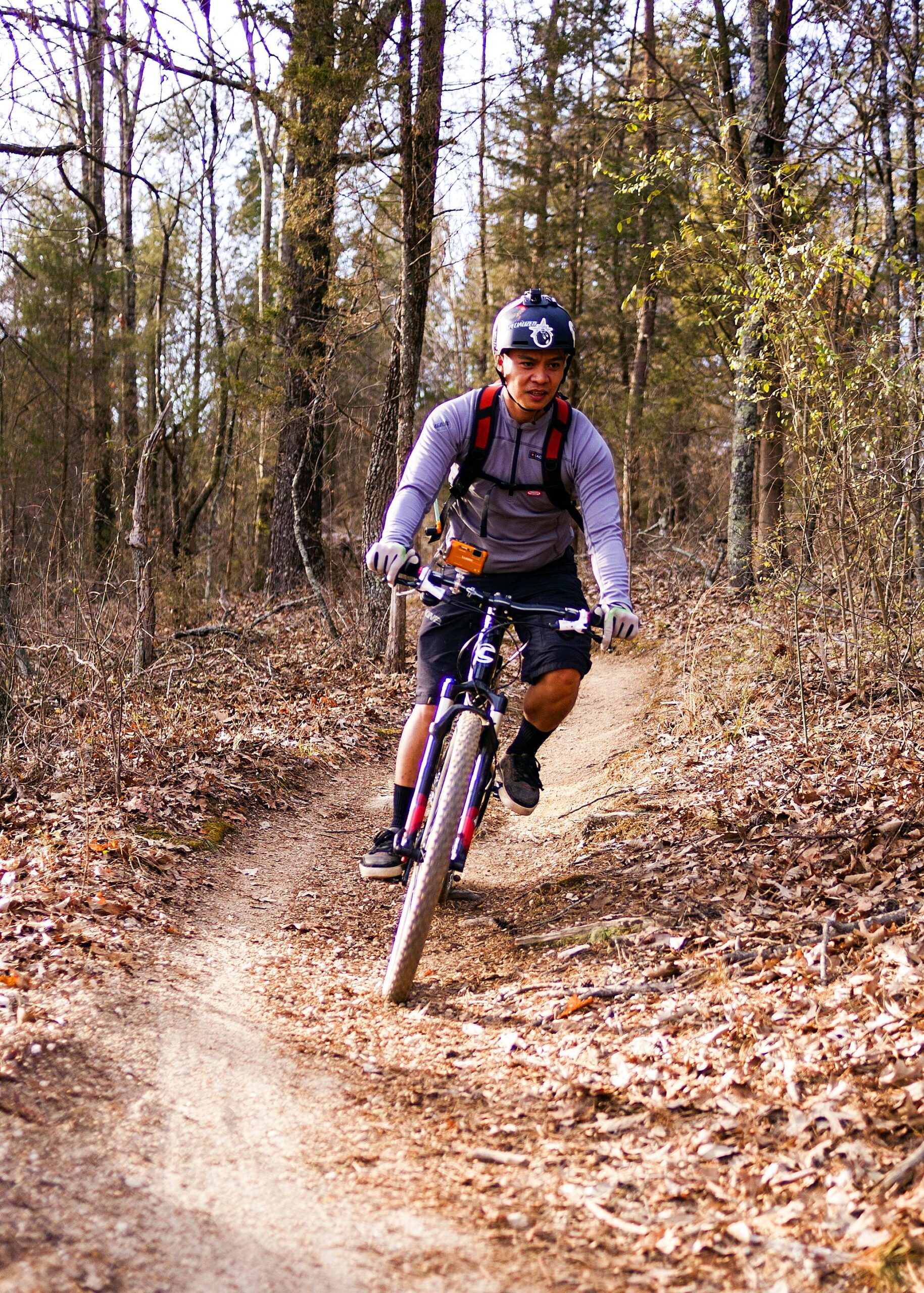 A person riding a mountain bike along a dirt trail in a wooded area, wearing a helmet and a backpack. The ground is covered in fallen leaves, and trees line the path. The rider appears focused and is navigating a turn in the trail. Hobby Park mountain bike trail.