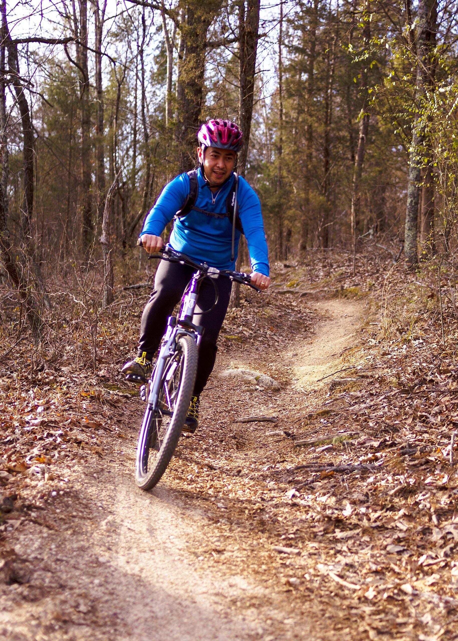 A person riding a mountain bike on a dirt trail surrounded by trees. The cyclist is wearing a pink helmet and a blue long-sleeve shirt, focused on the ride. The path is lined with fallen leaves and rocky terrain, suggesting an outdoor adventure. Hobby Park mountain bike trail.