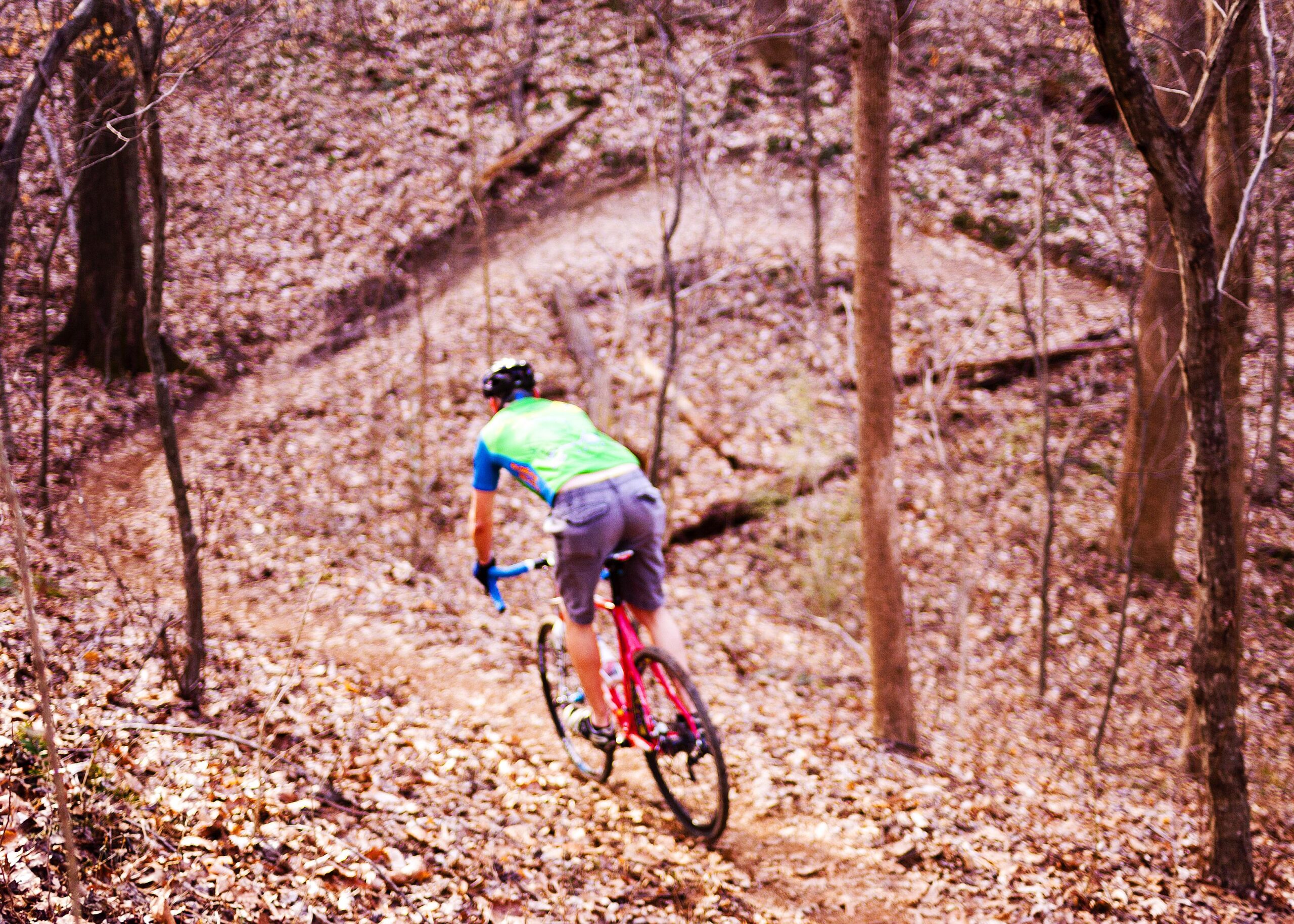 A cyclist wearing a bright green and blue jersey rides downhill on a dirt trail surrounded by autumn foliage. Trees and fallen leaves are visible in the background, creating a natural woodland setting. The image captures the dynamic motion of the cyclist as they navigate the winding path. Hobby Park mountain bike trail.