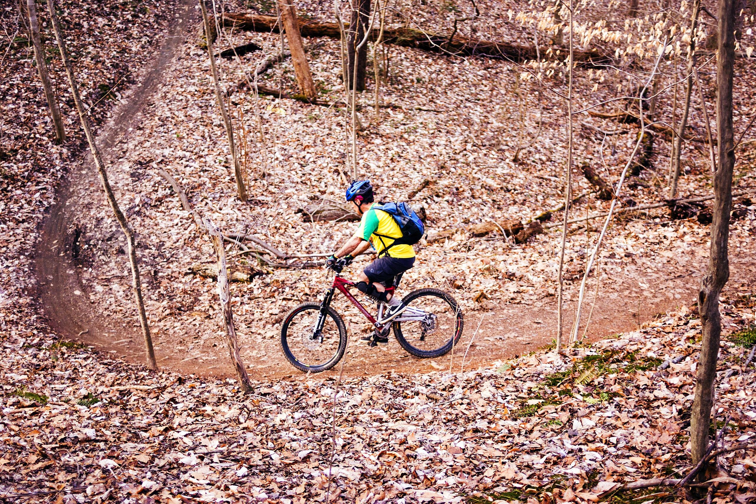 A mountain biker riding along a winding dirt trail surrounded by trees and fallen leaves in a forest setting. The cyclist is wearing a helmet and a colorful shirt, with a backpack on their back. Hobby Park mountain bike trail.