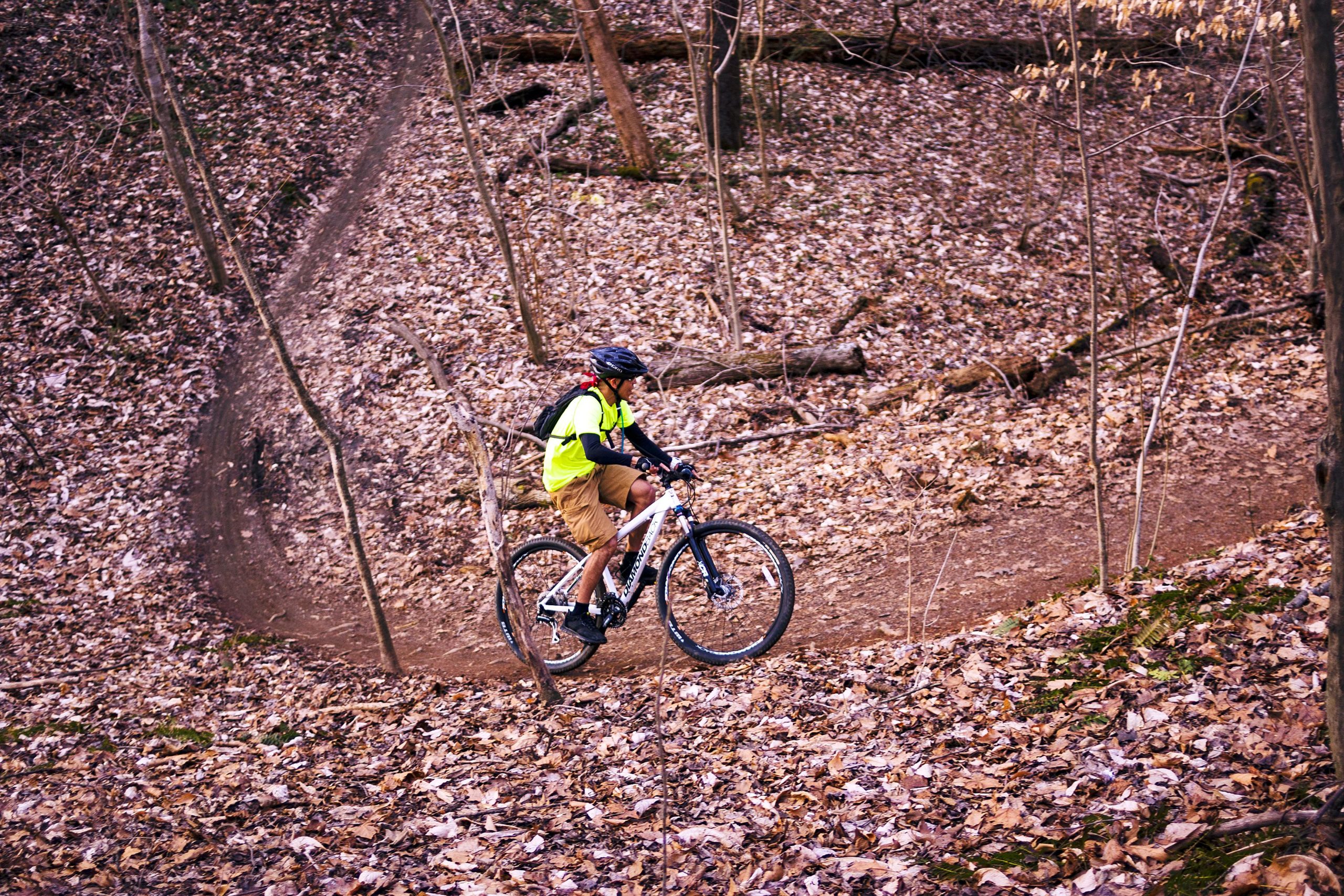 A mountain biker riding on a dirt trail covered with fallen leaves in a wooded area, wearing a bright yellow vest and a helmet. The bike is navigating a curved path surrounded by trees and underbrush. Hobby Park mountain bike trail.