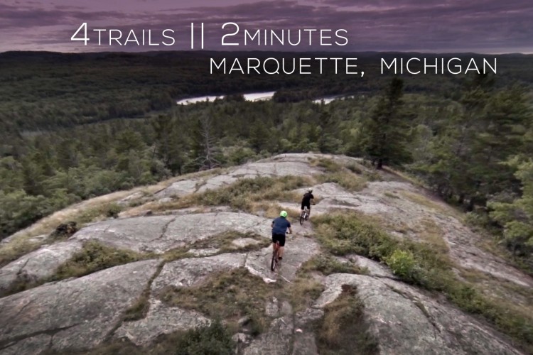 Aerial view of two mountain bikers navigating rocky trails in Marquette, Michigan, surrounded by lush greenery and a winding river in the background. Text overlay reads "4 TRAILS || 2 MINUTES."