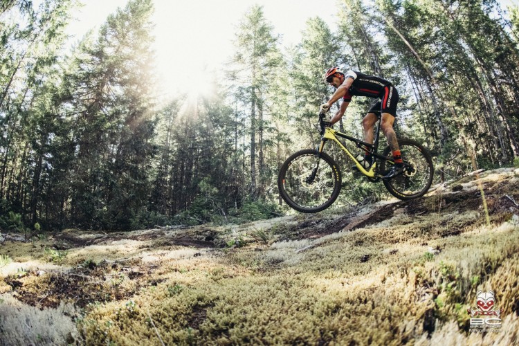 A mountain biker is riding down a trail in a dense forest, surrounded by tall trees and dappled sunlight filtering through the leaves. The cyclist is seen in a dynamic position, with one tire lifted off the ground as they navigate over a mossy terrain.